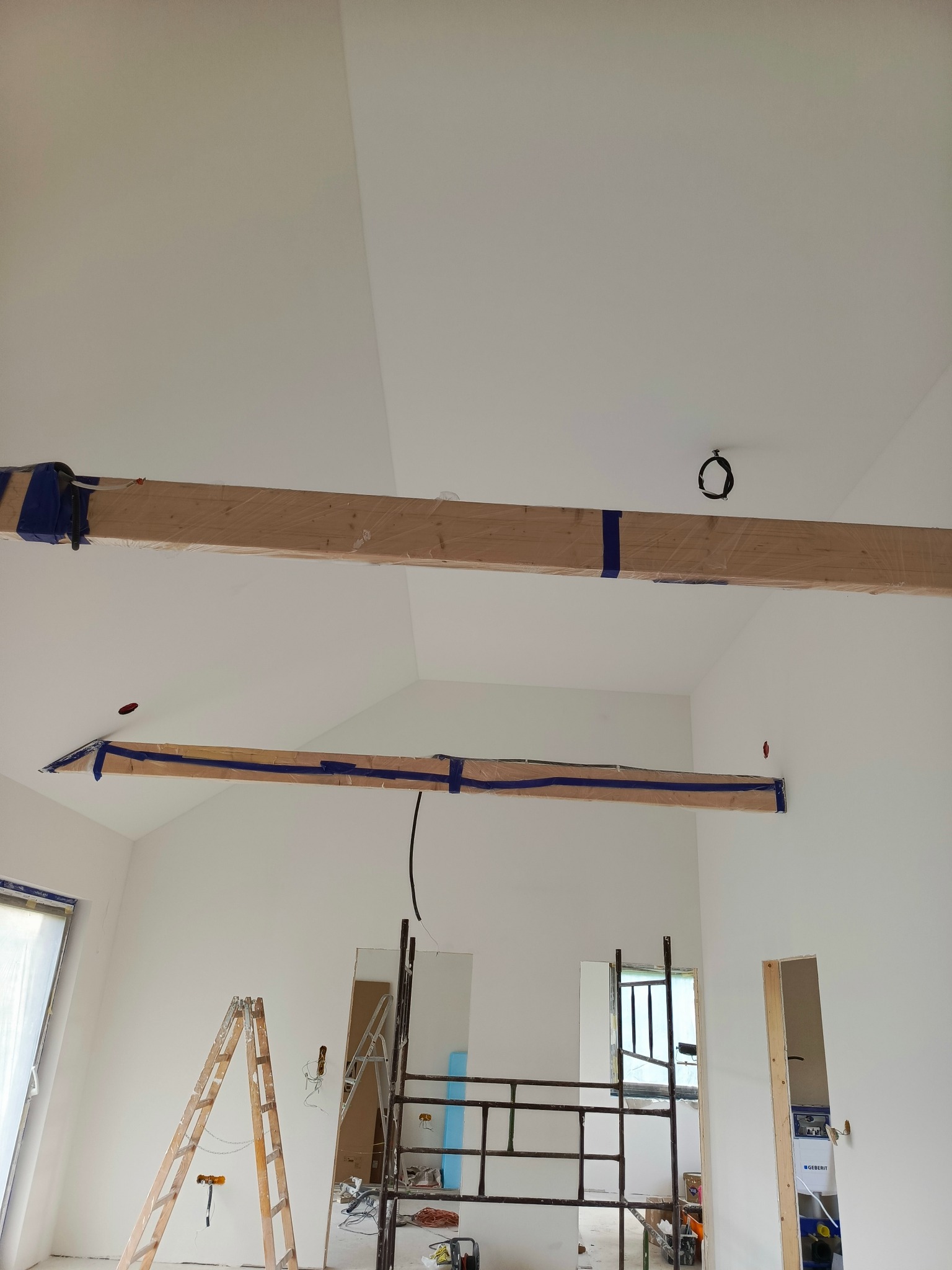 Wnętrze w trakcie prac wykończeniowych: widoczne drewniane belki na suficie zabezpieczone folią i taśmą malarską, drabiny i rusztowanie w pomieszczeniu z białymi ścianami.