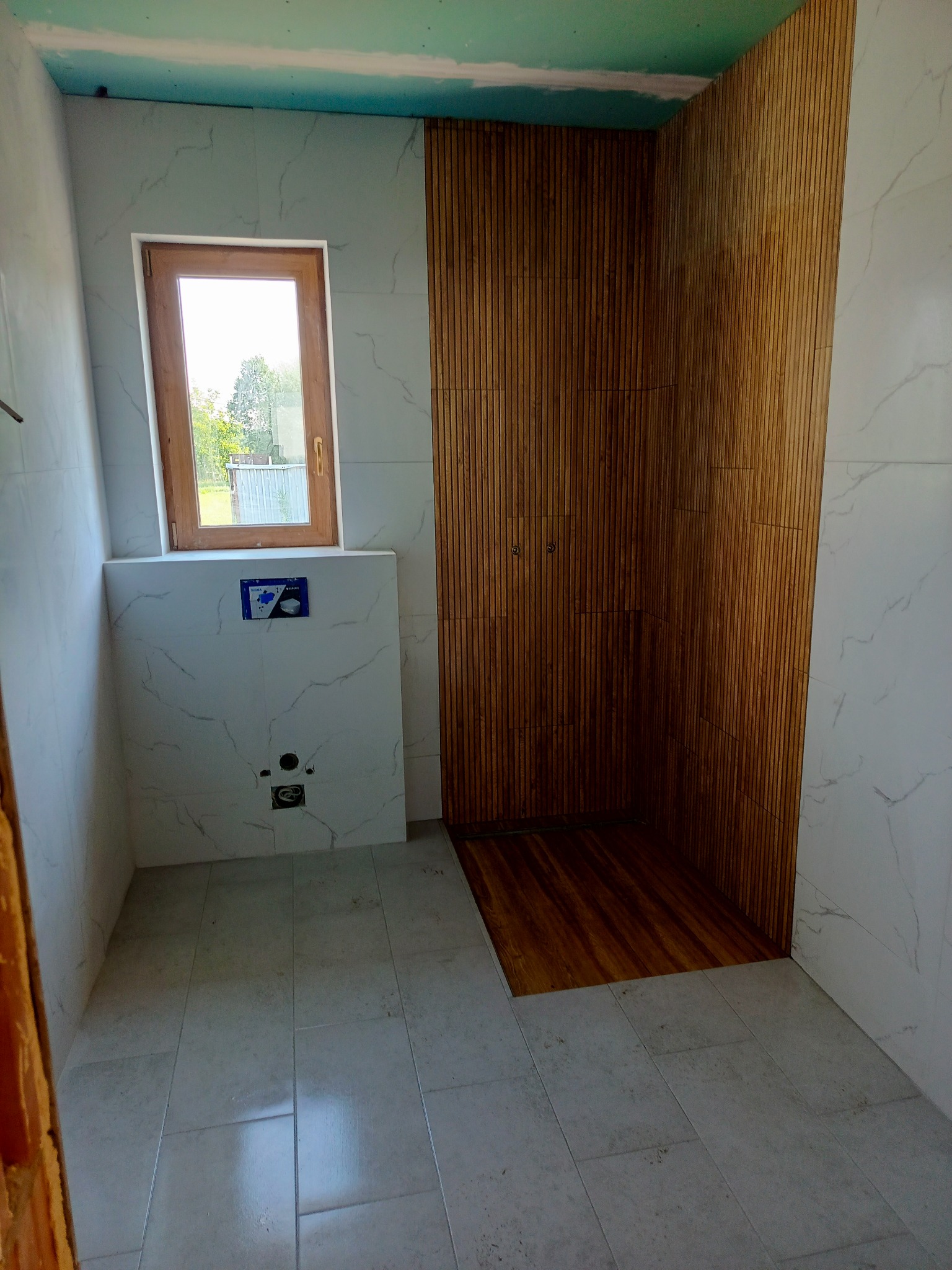 Wnętrze łazienki w trakcie wykańczania: białe płytki imitujące marmur, drewniana okładzina ścienna w strefie prysznica, surowy sufit z płyt kartonowo-gipsowych, oraz okno z widokiem na zieleń.