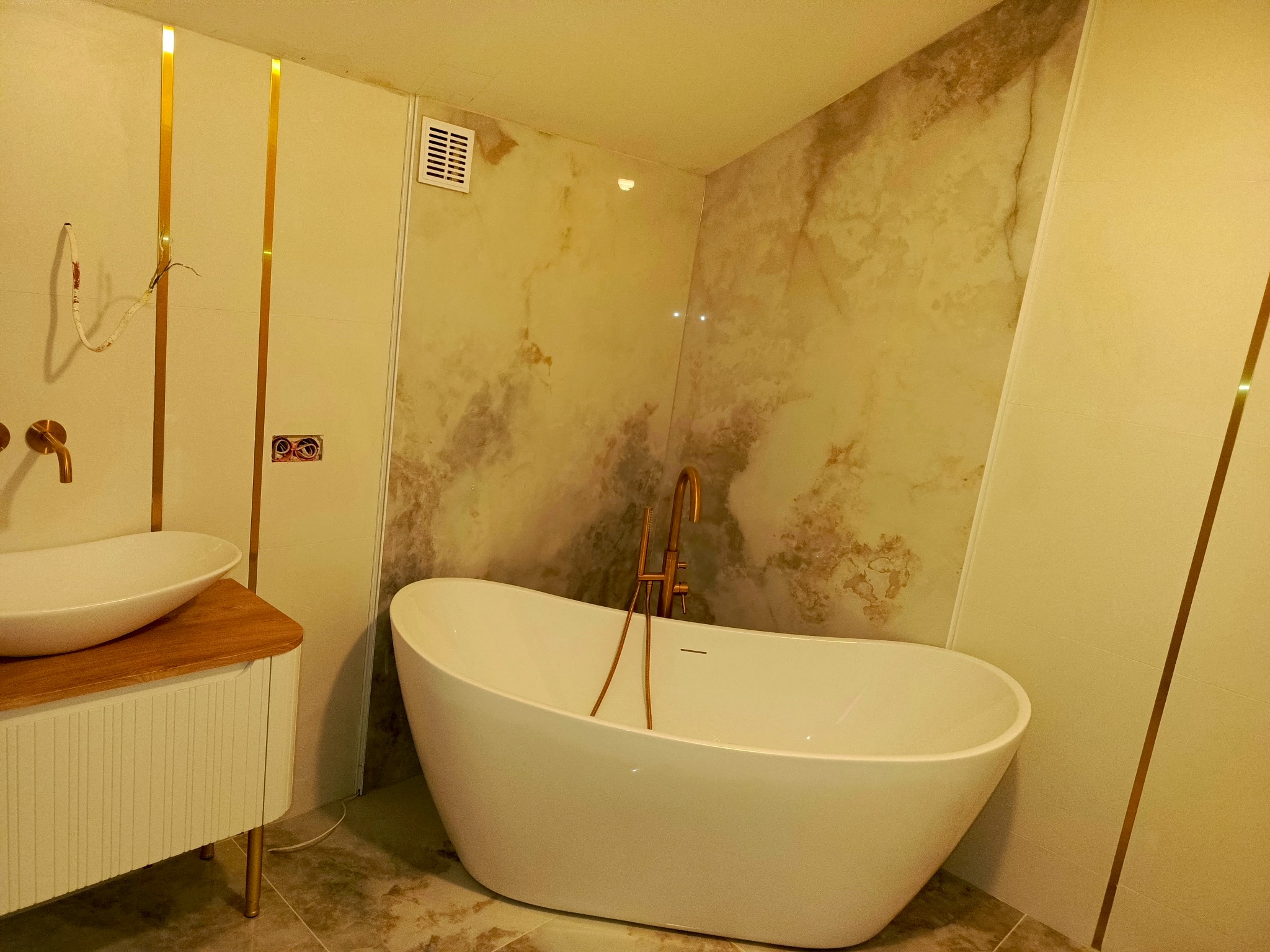 Nowoczesna łazienka z wolnostojącą białą wanną, umywalką nablatową na drewnianym blacie, marmurkowymi płytkami i złotymi akcentami.
