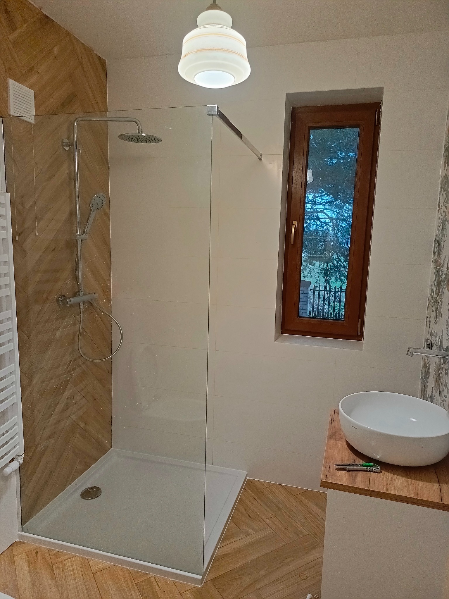 Nowoczesna łazienka z kabiną prysznicową typu walk-in, wykończona białymi i drewnopodobnymi płytkami, z umywalką nablatową i oknem z widokiem na zieleń.