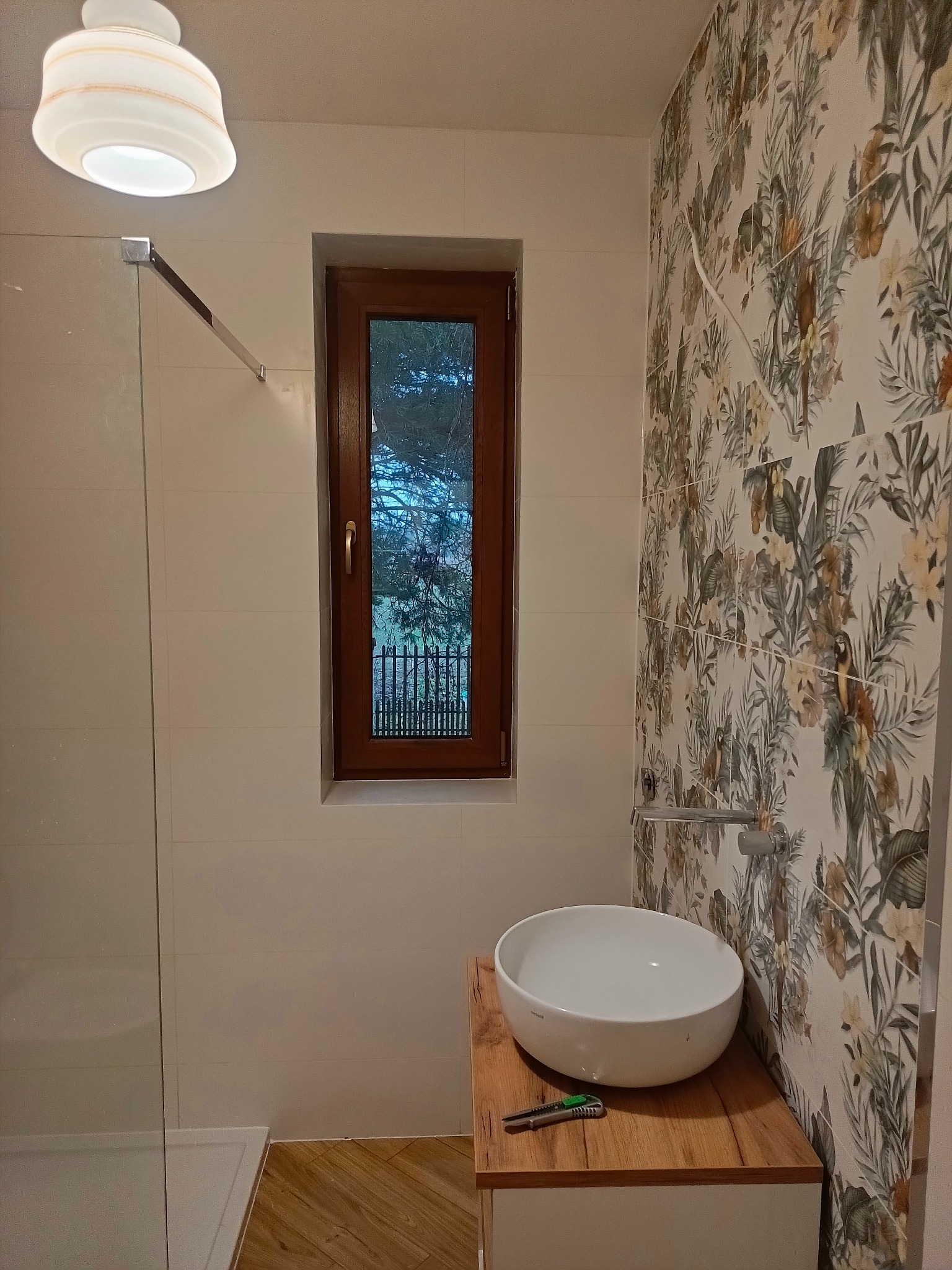 Nowoczesna łazienka po remoncie: biała umywalka nablatowa na drewnianym blacie, ściany wyłożone płytkami z motywem tropikalnym, okno z widokiem na zieleń, kabina prysznicowa w tle, nóż...