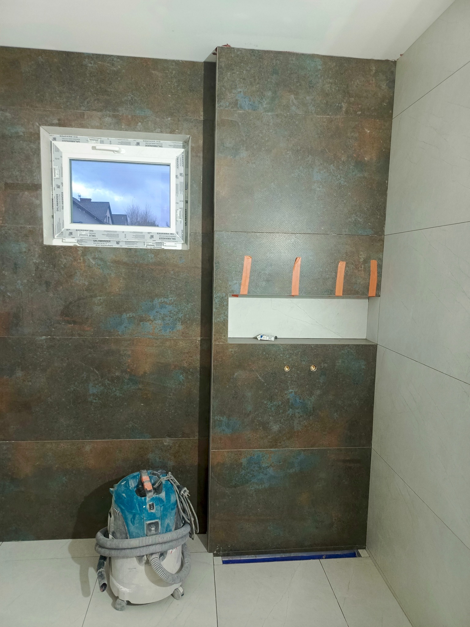 Łazienka w trakcie wykańczania: ściany pokryte mozaiką w odcieniach brązu i błękitu, wnęka z białymi płytkami zabezpieczona pomarańczową taśmą, okno z widokiem na dom, odkurzacz przemysłowy...