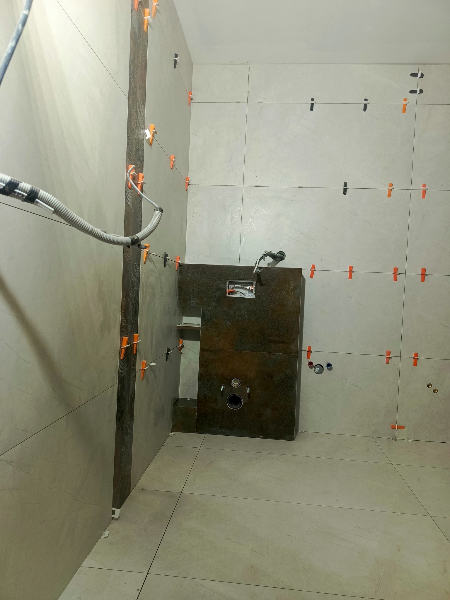 Łazienka w trakcie układania płytek, widoczne krzyżyki dystansowe i pozioma listwa dekoracyjna z ciemnych płytek, instalacja podtynkowa WC w zabudowie.