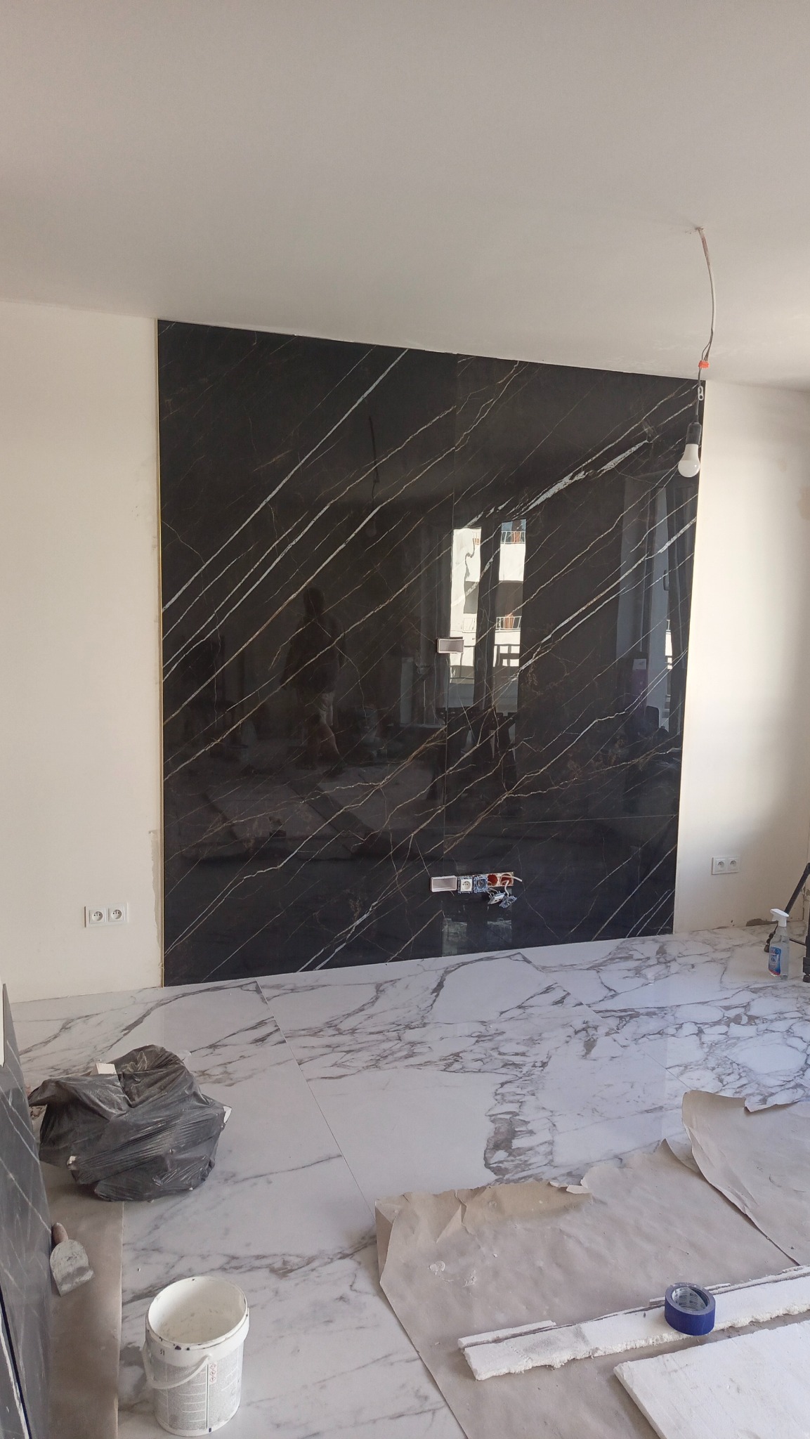 Ściana wyłożona czarnymi płytami z białym marmurkowym wzorem, dopasowana do marmurowej podłogi, widoczne gniazdka elektryczne, kabel z żarówką i narzędzia glazurnicze