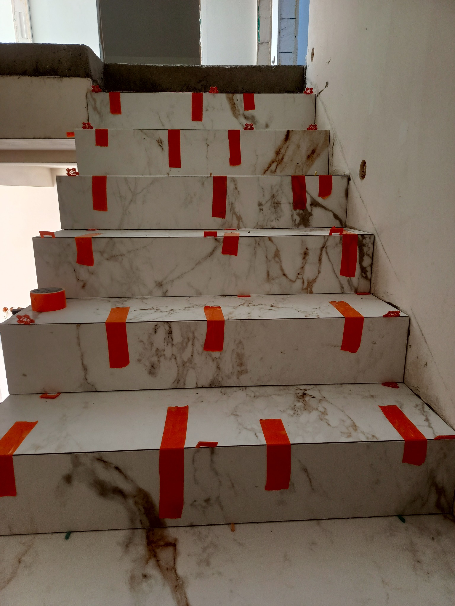 Schody wyłożone płytkami imitującymi marmur w trakcie montażu, zabezpieczone taśmą i poziomowane krzyżykami. Widoczne surowe ściany i niedokończone elementy.