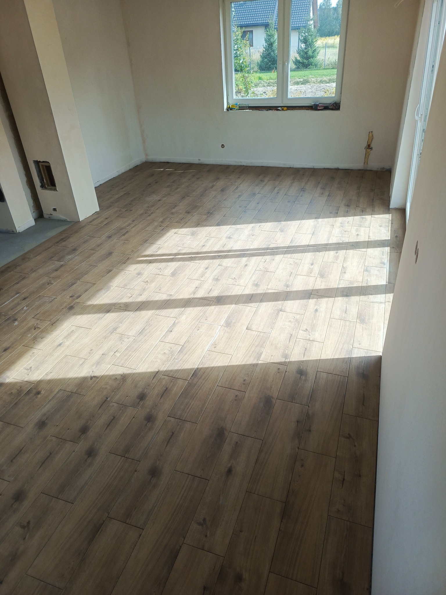 Ukończona podłoga z płytek imitujących drewno w jasnym, nowym pomieszczeniu z oknem. Płytki ułożone w regularny wzór, widoczne promienie słoneczne.