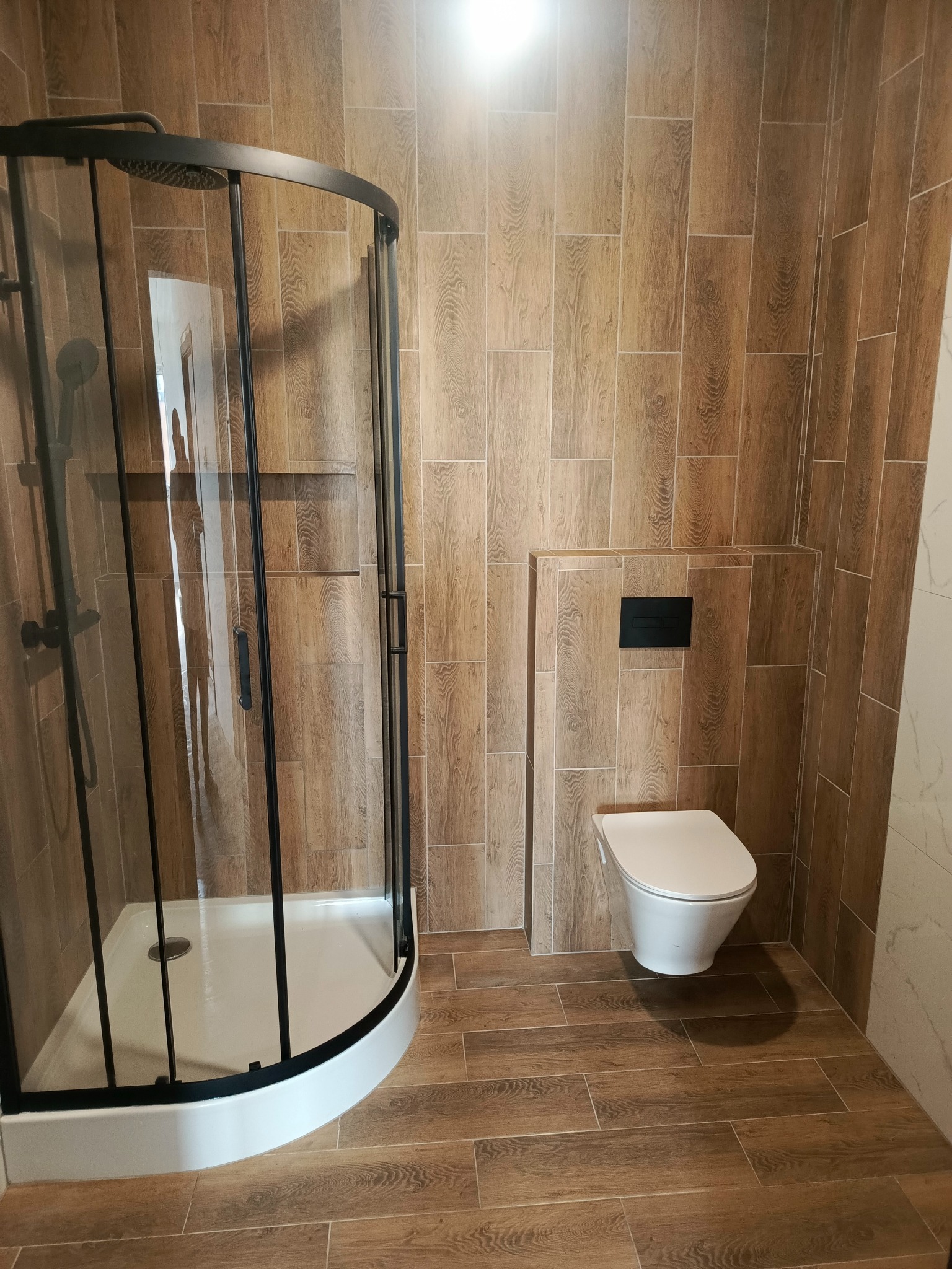 Wykończona łazienka z kabiną prysznicową typu walk-in z czarnymi ramami i toaletą podwieszaną. Płytki imitujące drewno na ścianach i podłodze.