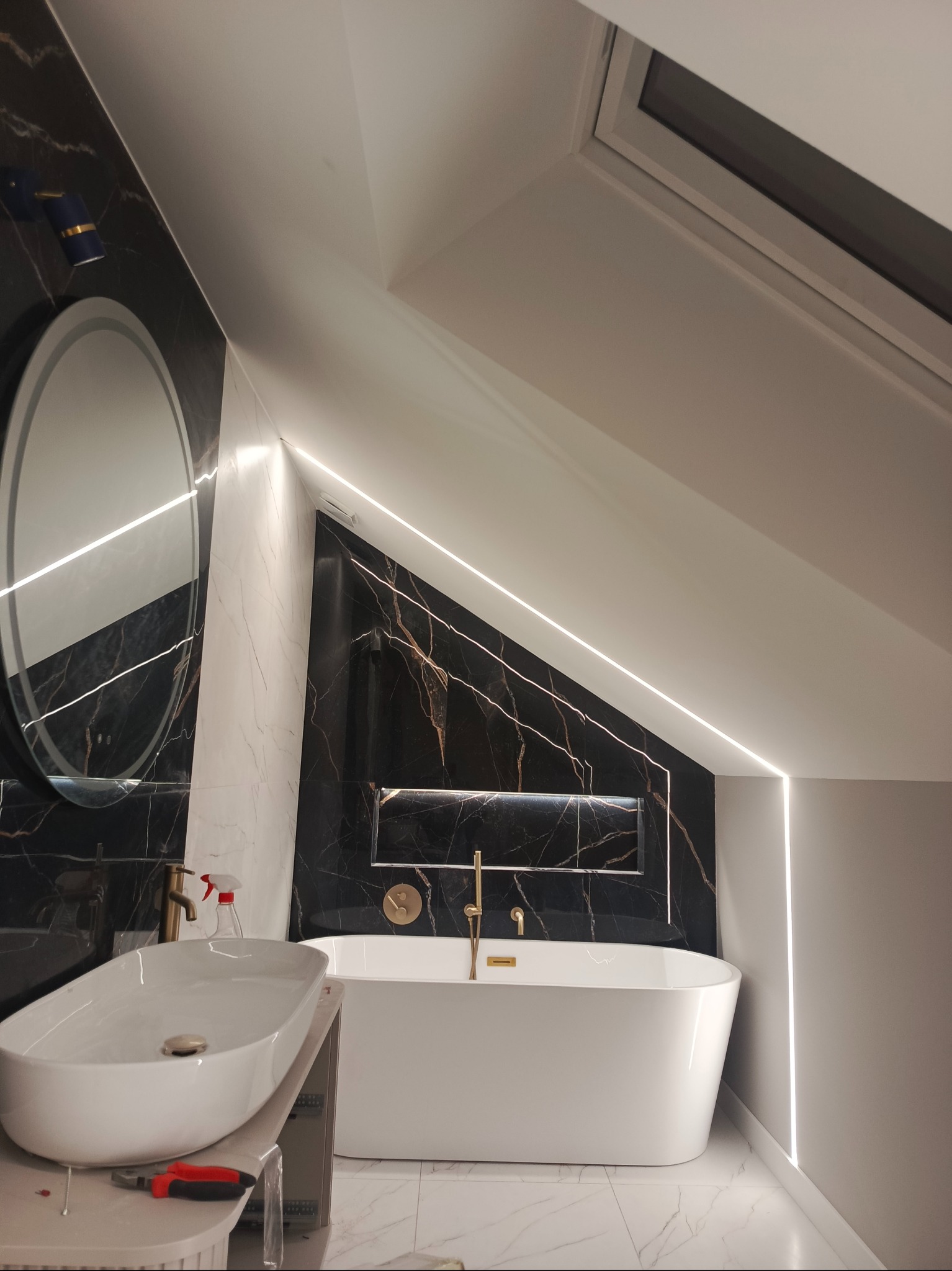 Nowoczesna łazienka na poddaszu z wanną wolnostojącą, umywalką nablatową i oświetleniem LED. Czarno-białe marmurowe płytki i złota armatura dodają elegancji.