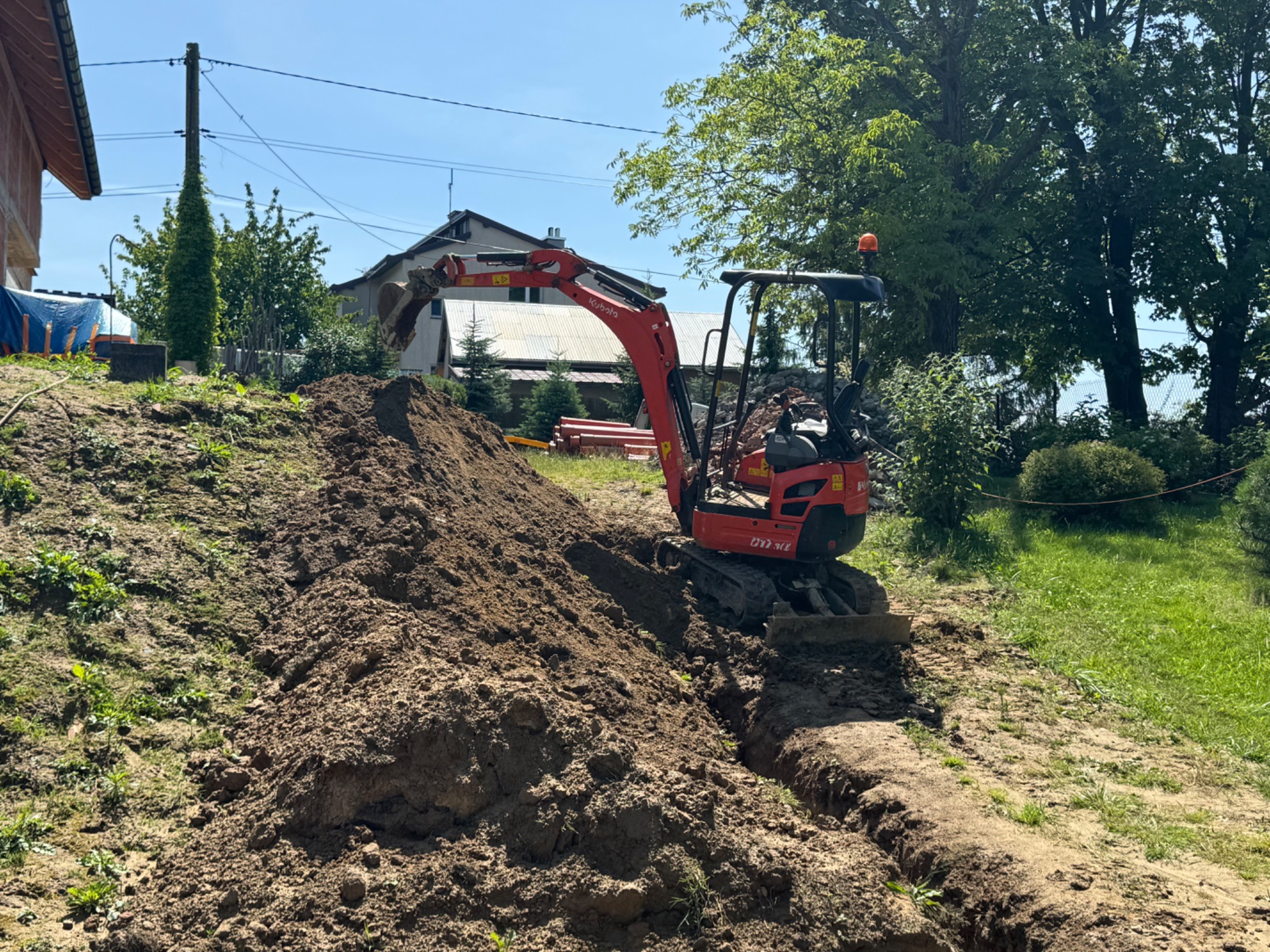 Czerwona mini koparka Kubota wykonuje wykop w ziemi na nierównym terenie z domem w tle.