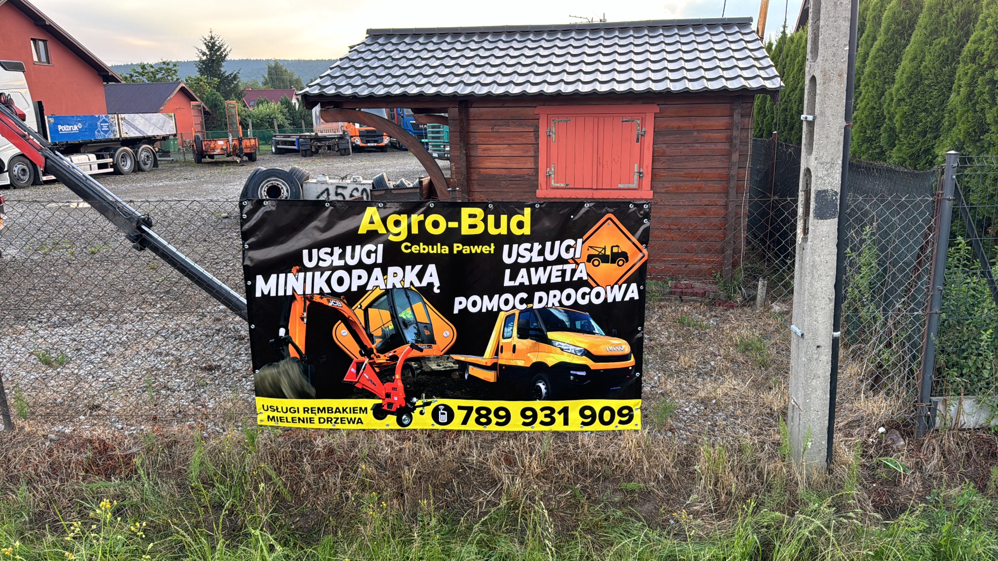 Baner reklamowy firmy Agro-Bud z usługami minikoparką, lawetą i rębakiem do mielenia drzewa, zawieszony na ogrodzeniu z siatki, w tle budynek gospodarczy i zaparkowane pojazdy.