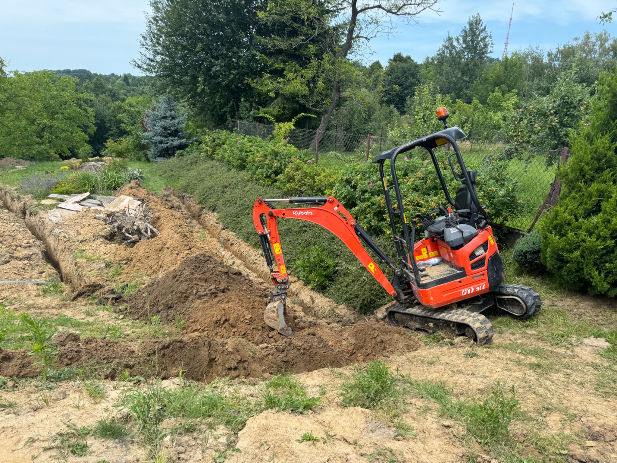 Pomarańczowa mini koparka Kubota wykonuje wykop liniowy w ogrodzie, widoczne ślady świeżej ziemi i zielona roślinność w tle.