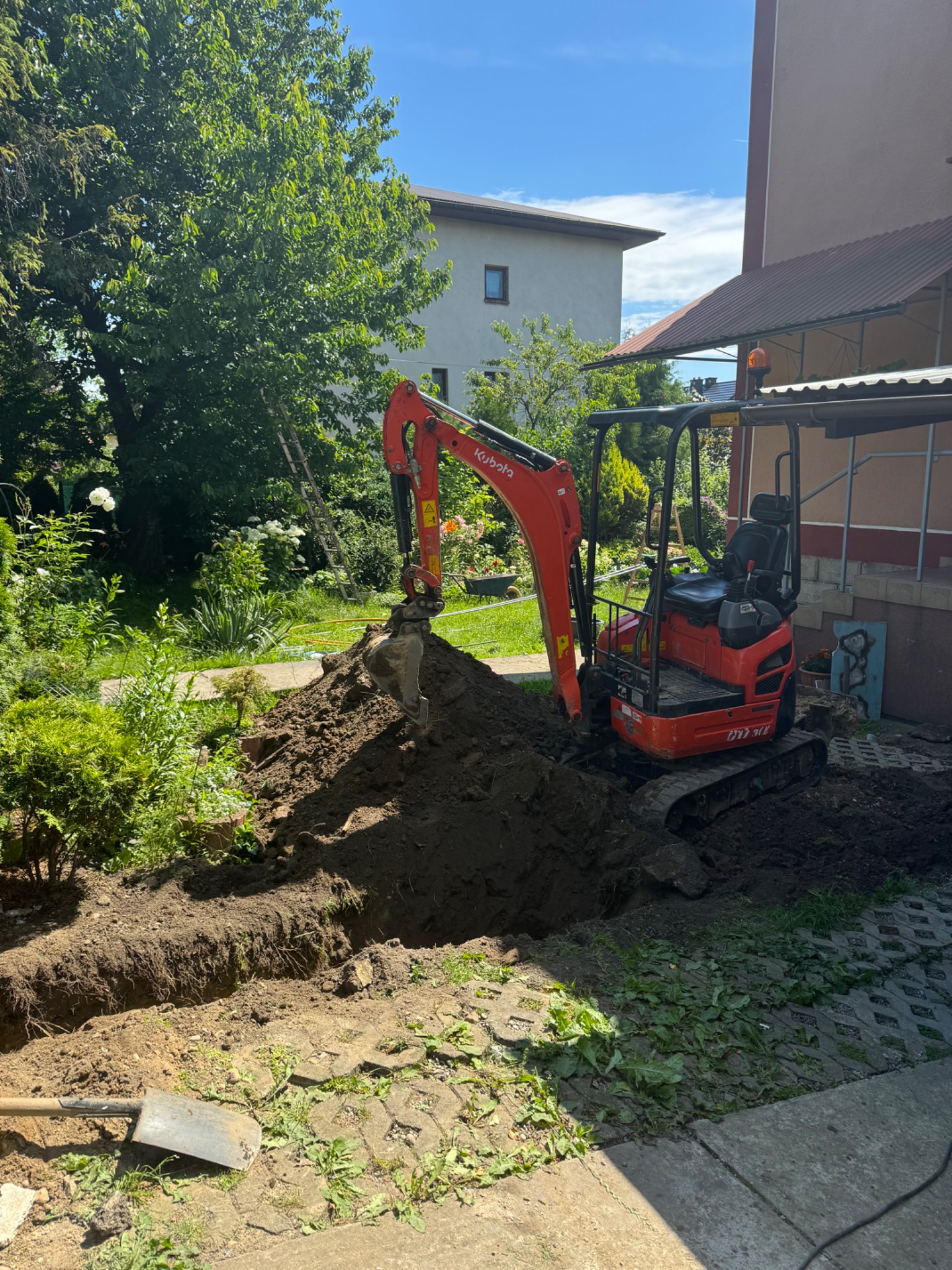 Czerwona mini koparka Kubota w trakcie prac ziemnych: wykop głębokiego rowu w ogrodzie, widoczna sterta ziemi, łopata i kostka brukowa w tle drzewa i budynek.