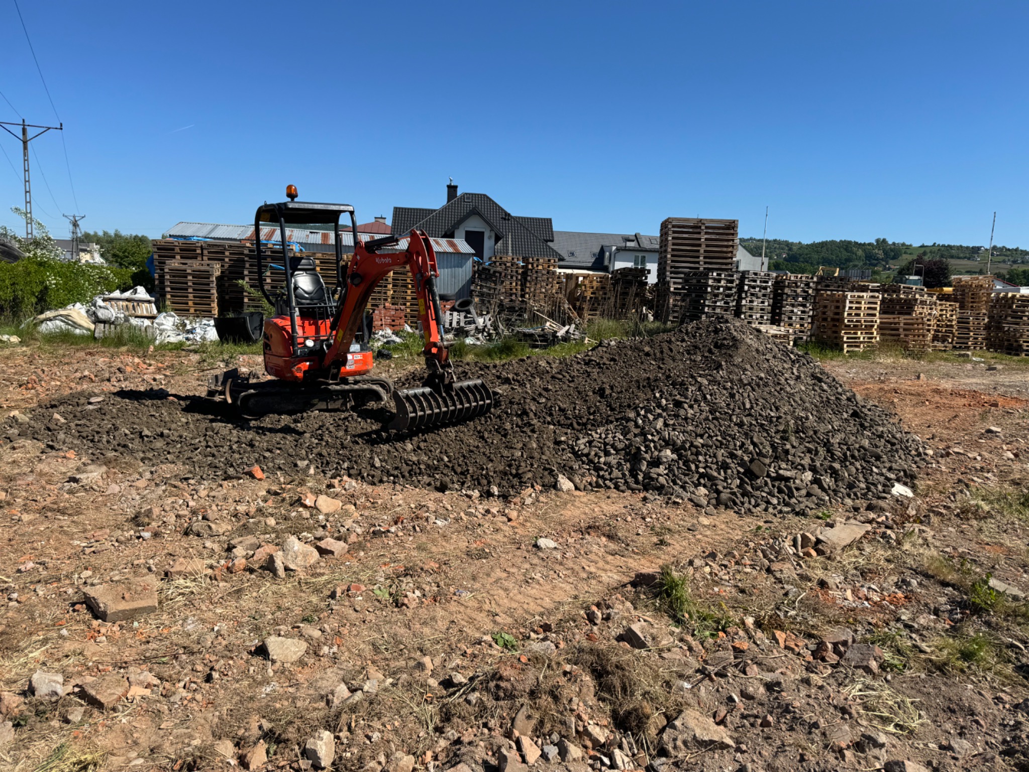 Pomarańczowa mini-koparka Kubota wyrównuje teren z pryzmą ziemi na działce, w tle sterty drewnianych palet i zabudowania.