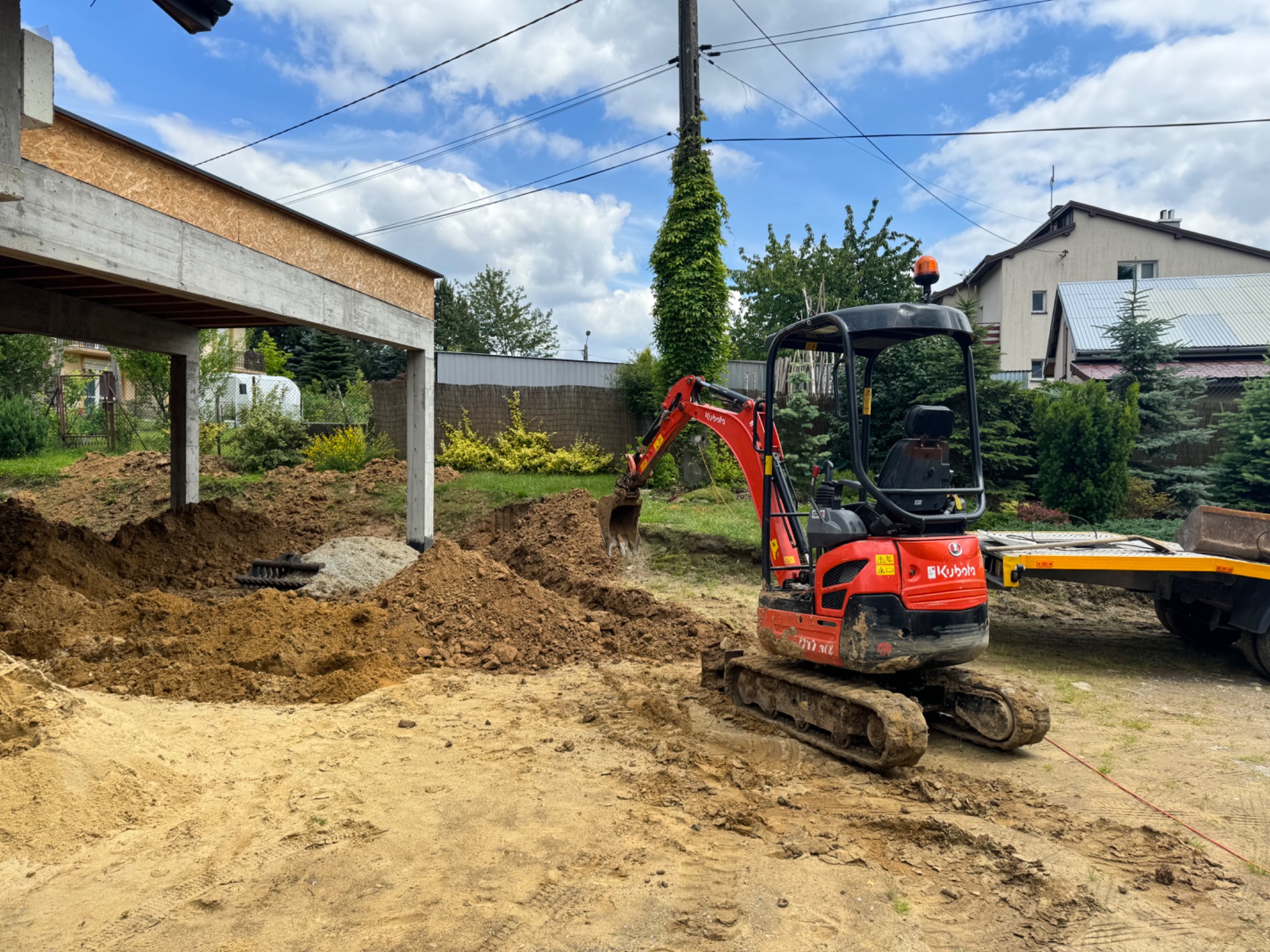 Czerwona mini koparka Kubota na placu budowy wykopuje ziemię pod fundamenty nowego budynku z betonowymi słupami, widoczna laweta i dom w tle.