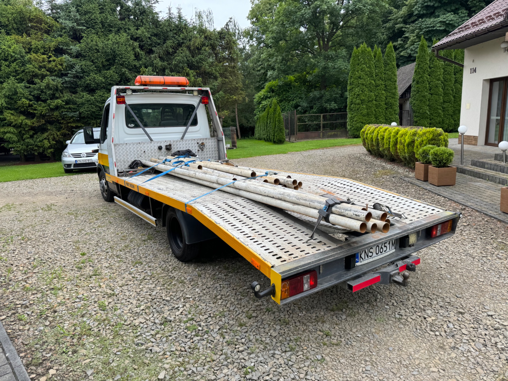 Biała laweta z pomarańczowym światłem ostrzegawczym przewozi rury zabezpieczone niebieskimi pasami, zaparkowana na żwirowym podjeździe przed domem.