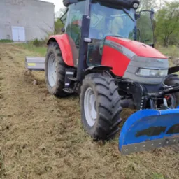 Czerwony ciągnik McCormick CX90 z kosiarką listwową i niebieskim pługiem na polu częściowo skoszonej trawy, widoczny budynek gospodarczy w tle.