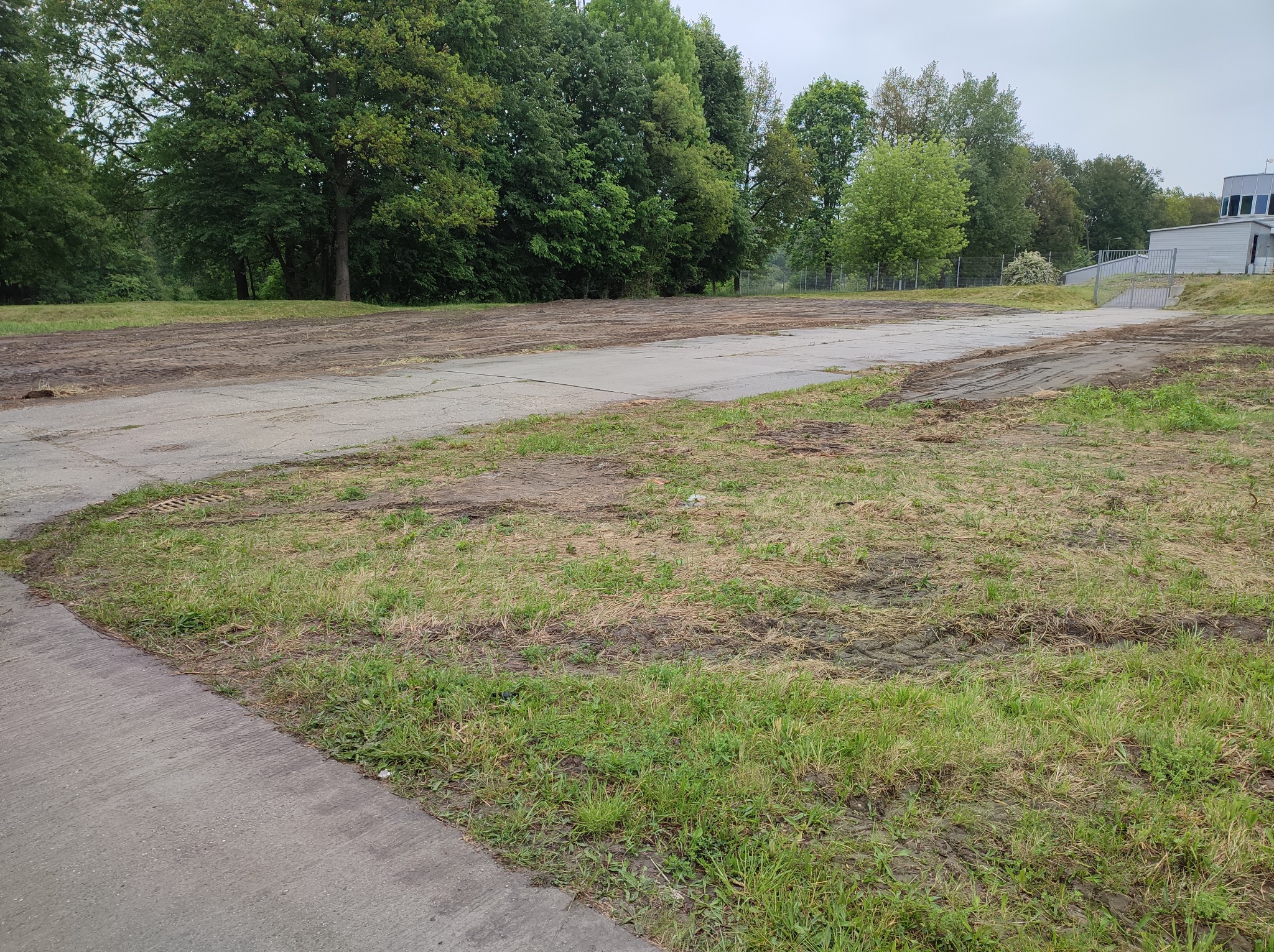 Przygotowany teren pod budowę z częściowo usuniętą warstwą humusu, widoczne ślady opon, w tle drzewa i budynek.