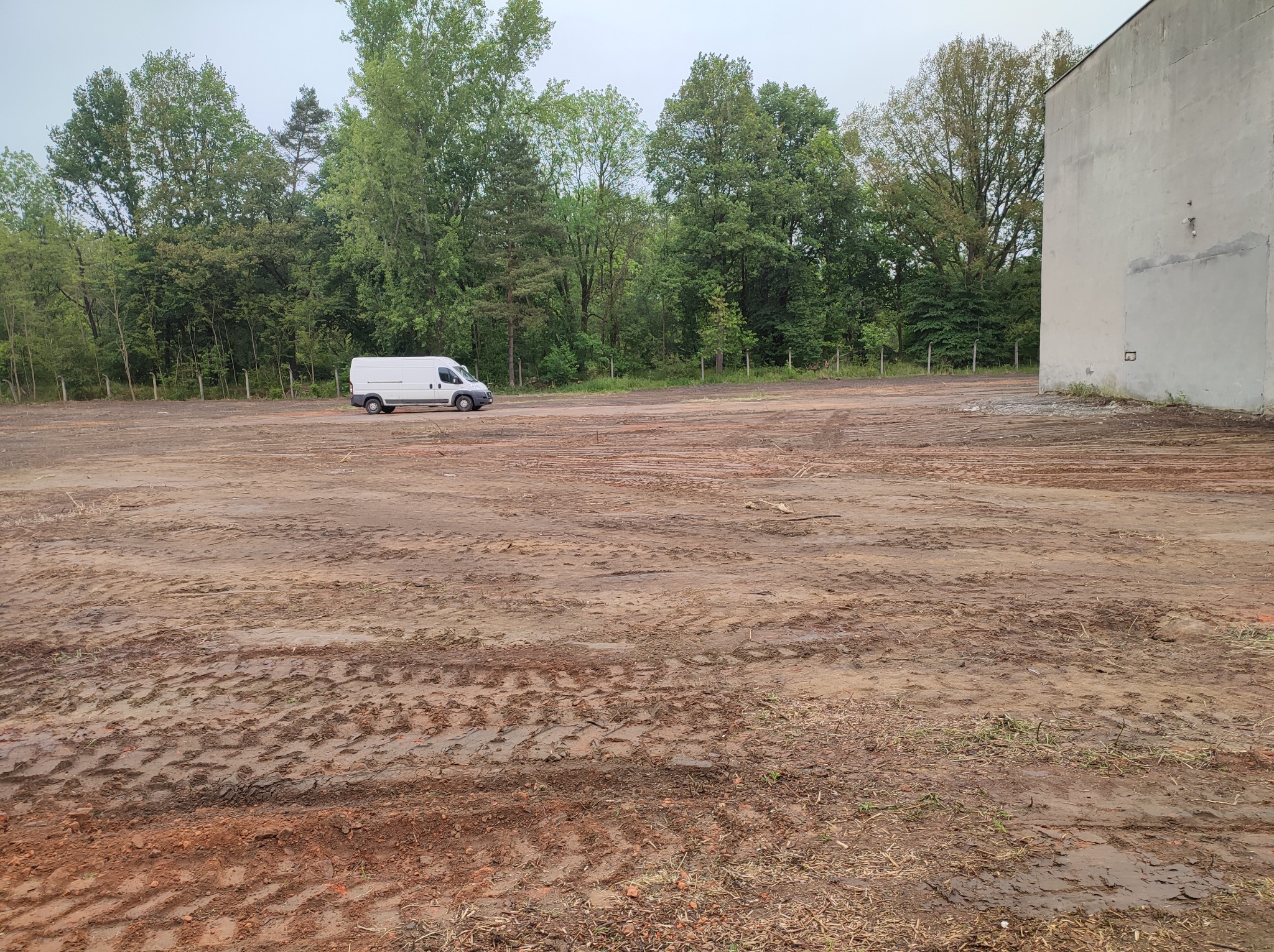 Biały bus na placu budowy z wyjeżdżoną, błotnistą ziemią, w tle drzewa i szary budynek.