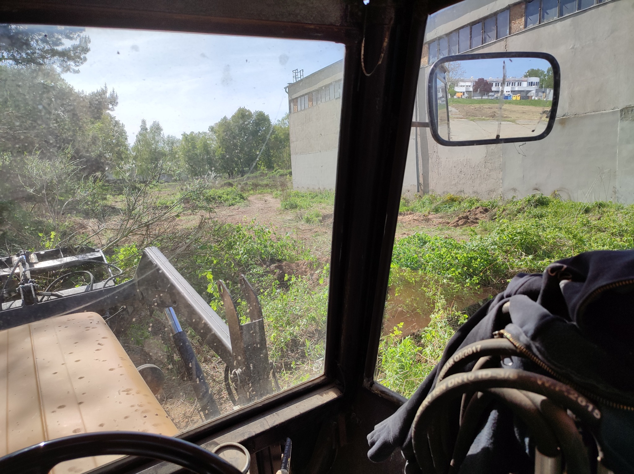 Widok z kabiny koparki na teren z częściowo usuniętą roślinnością, budynek w tle i odbicie w lusterku
