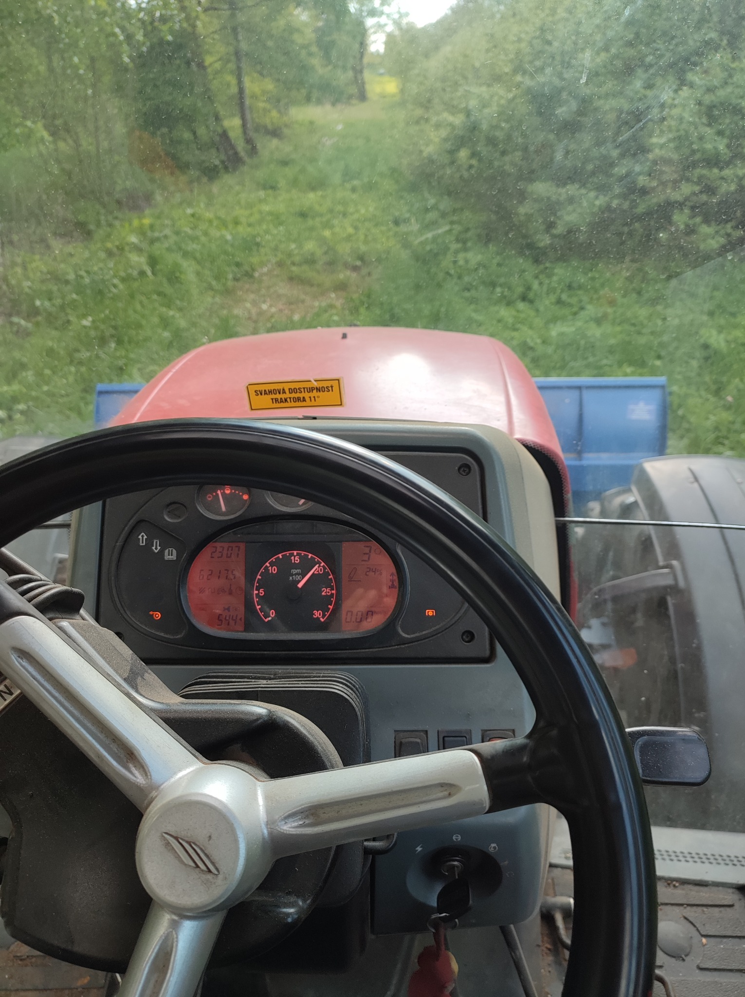 Widok z kabiny traktora na zewnątrz, z widocznymi wskaźnikami na desce rozdzielczej i fragmentem kierownicy. Na zewnątrz zieleń i drzewa.