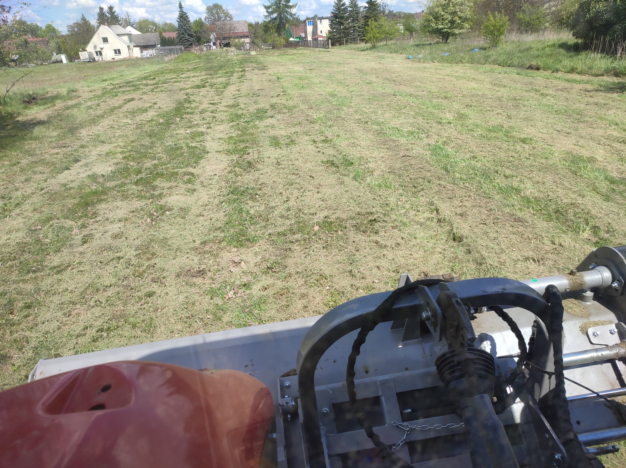 Widok z perspektywy operatora na rozległy teren po skoszeniu, z widocznymi pasami świeżo ściętej trawy i fragmentami maszyny koszącej w dolnej części kadru, w tle zabudowa jednorodzinna i drzewa.