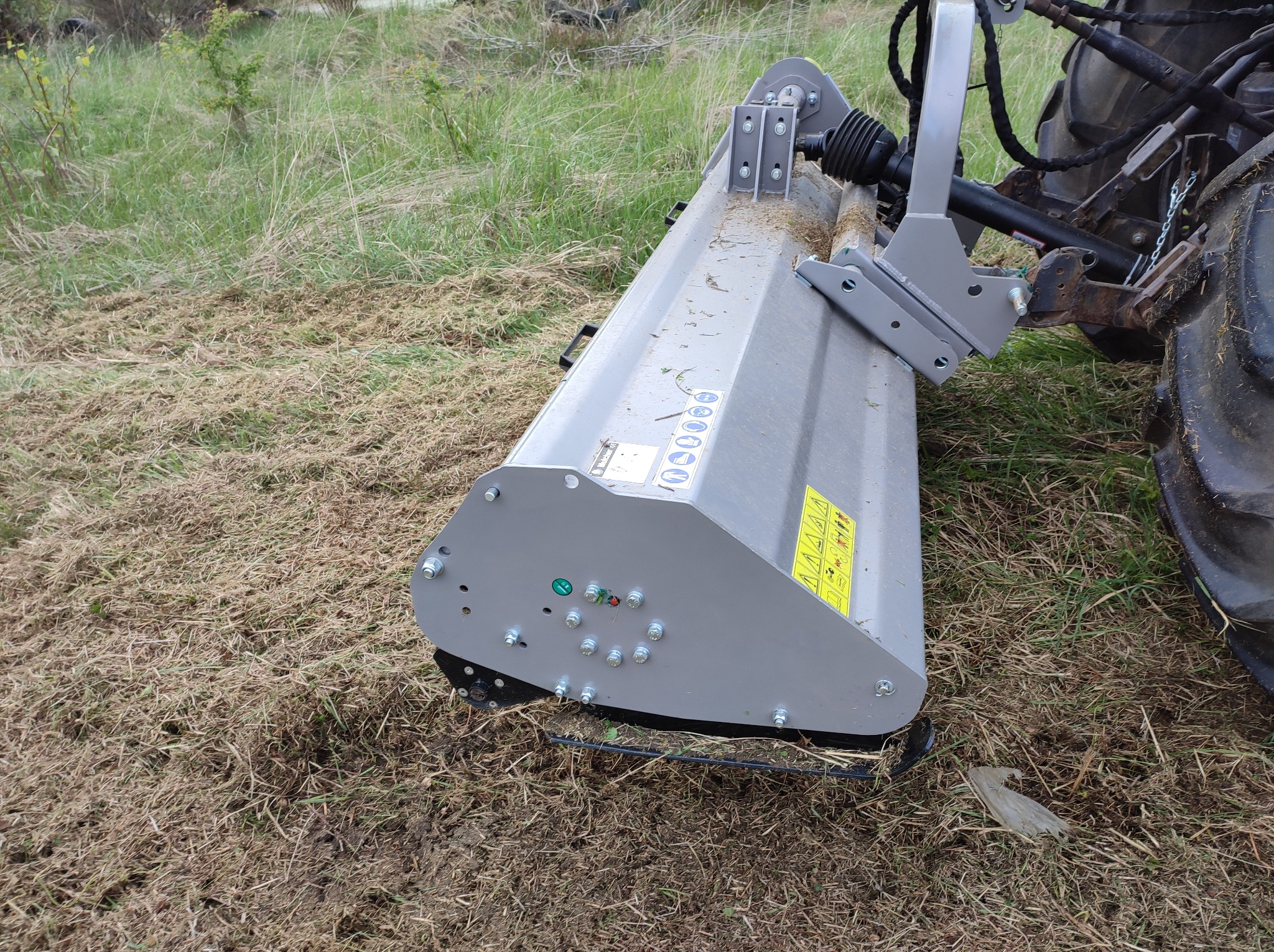 Głowica kosiarki bijakowej zamontowana na ciągniku, widoczna po koszeniu trawy, z żółtymi naklejkami ostrzegawczymi i zielonymi wskaźnikami.