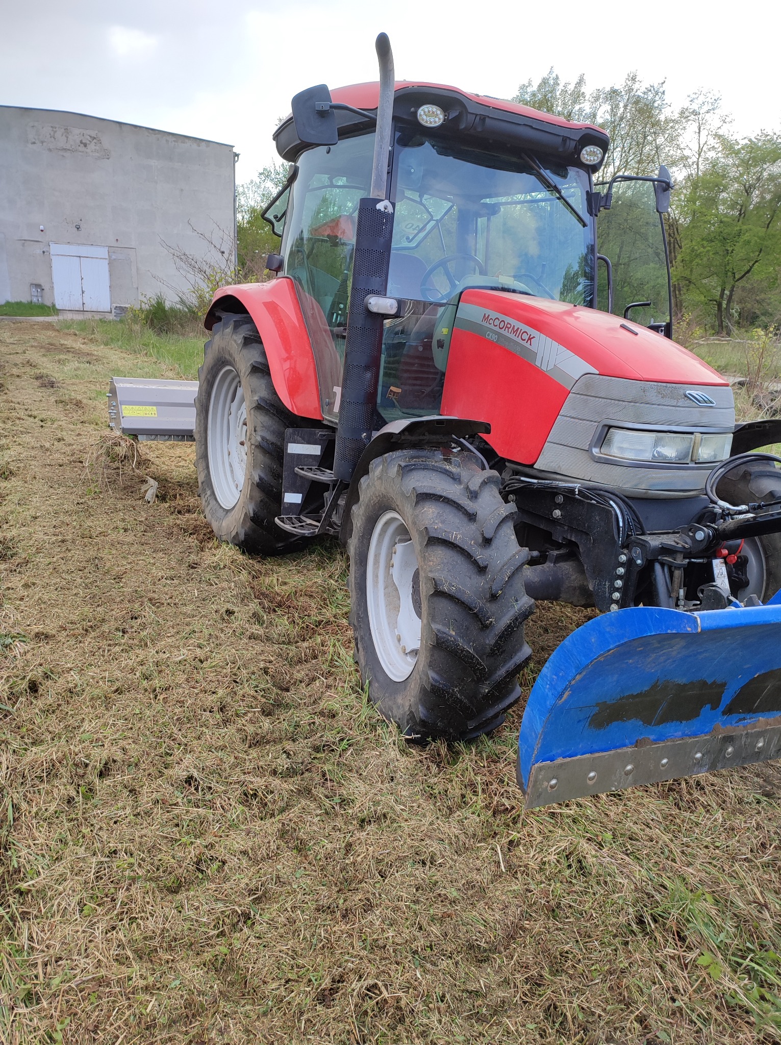 Czerwony ciągnik McCormick CX90 z kosiarką listwową i niebieskim pługiem na polu częściowo skoszonej trawy, widoczny budynek gospodarczy w tle.