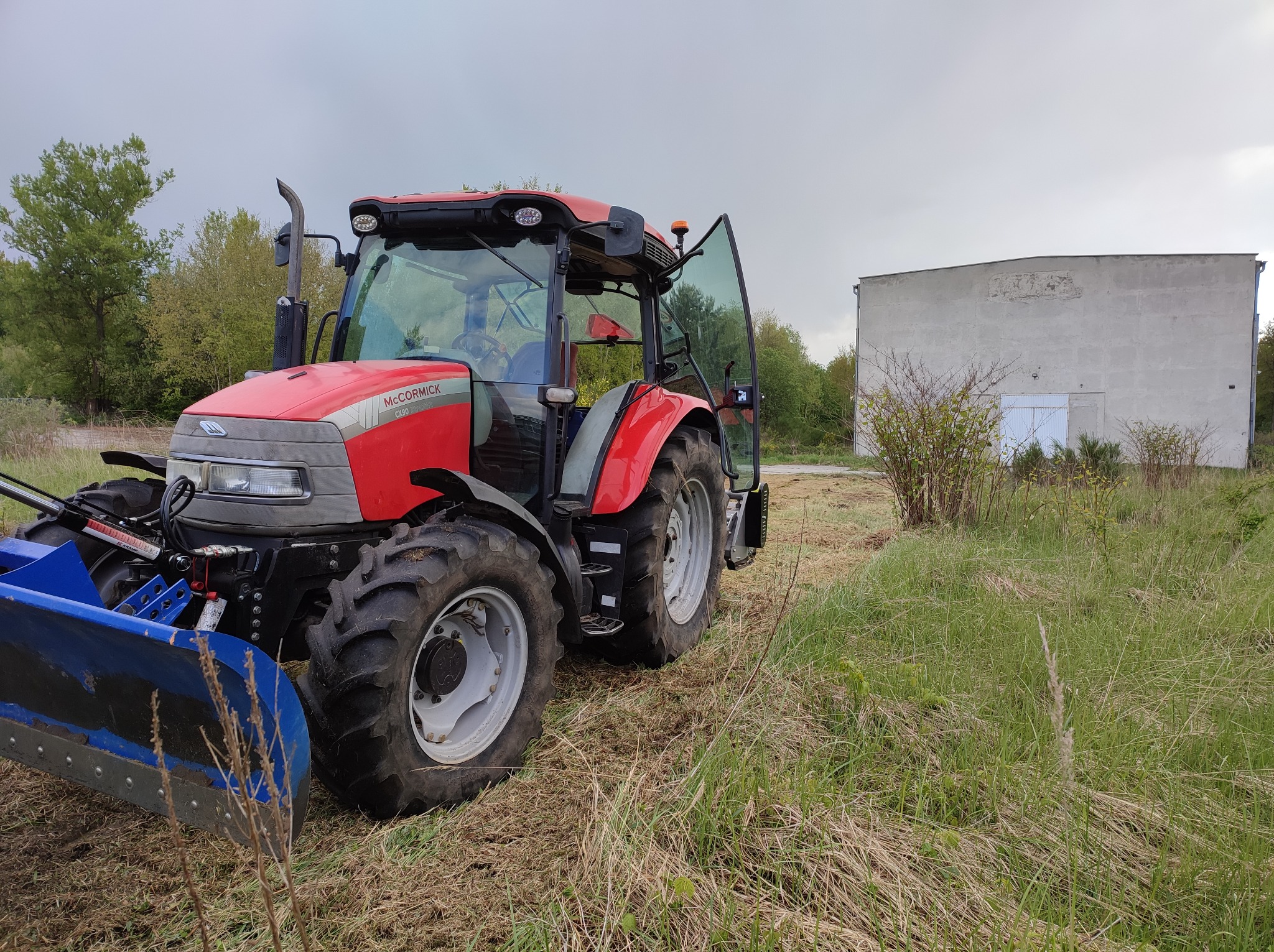 Czerwony traktor McCormick z otwartymi drzwiami, z zamontowanym niebieskim pługiem, stoi na łące z budynkiem w tle, w pochmurny dzień.