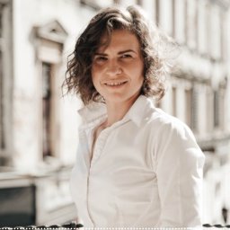 EUROVERA GROUP Ewelina Gryczka - Portret uśmiechniętej kobiety w białej koszuli na tle jasnej architektury z balkonem, z lekkim efektem vintage.