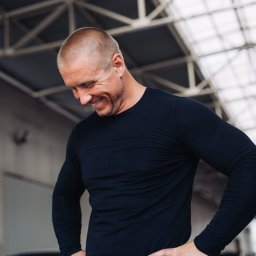 Mgr Adrian Markowicz - Uśmiechnięty mężczyzna w czarnej koszulce i jeansach, z rękami na biodrach, spogląda w dół. W tle konstrukcja dachu i zaparkowane samochody.