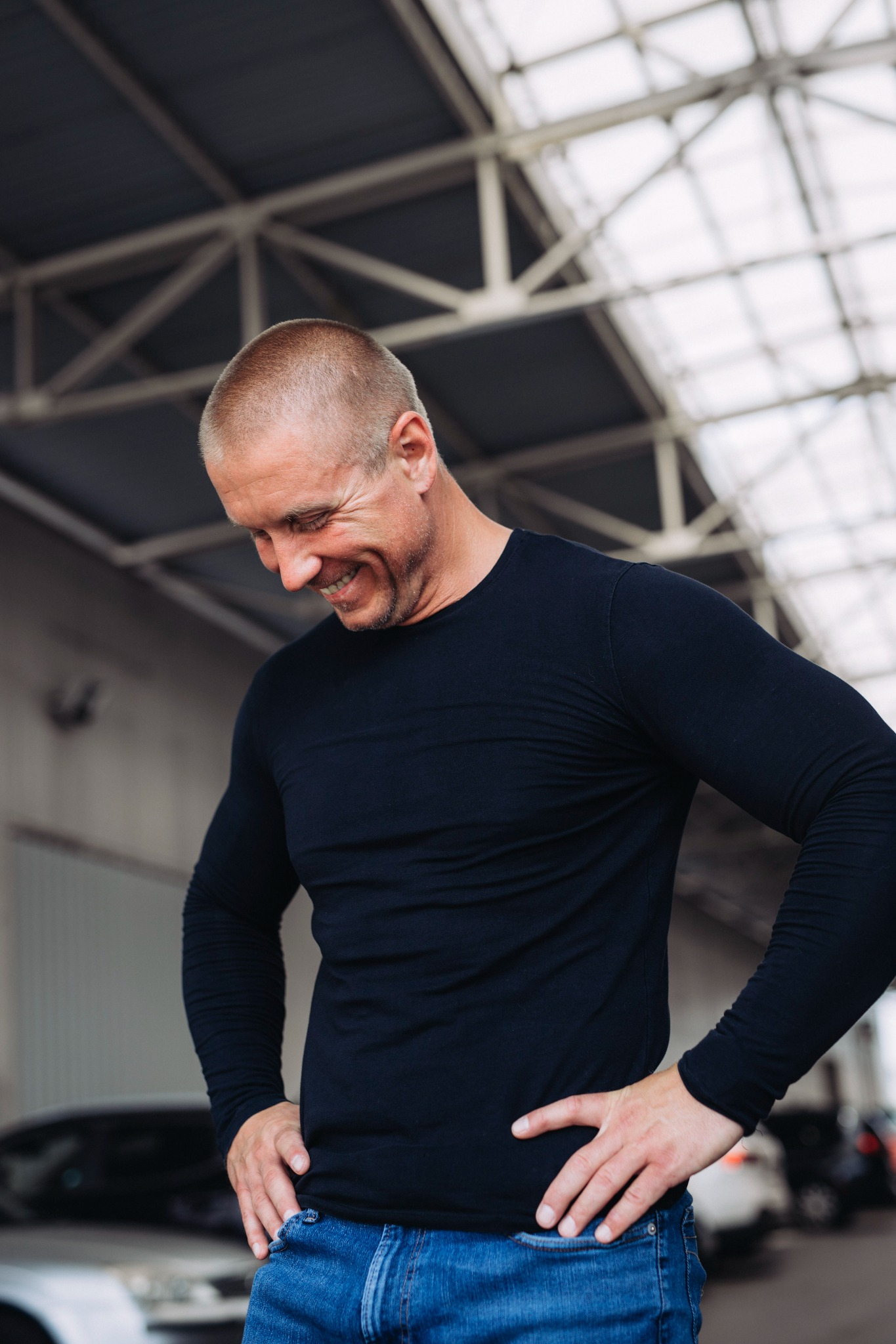 Uśmiechnięty mężczyzna w czarnej koszulce i jeansach, z rękami na biodrach, spogląda w dół. W tle konstrukcja dachu i zaparkowane samochody.