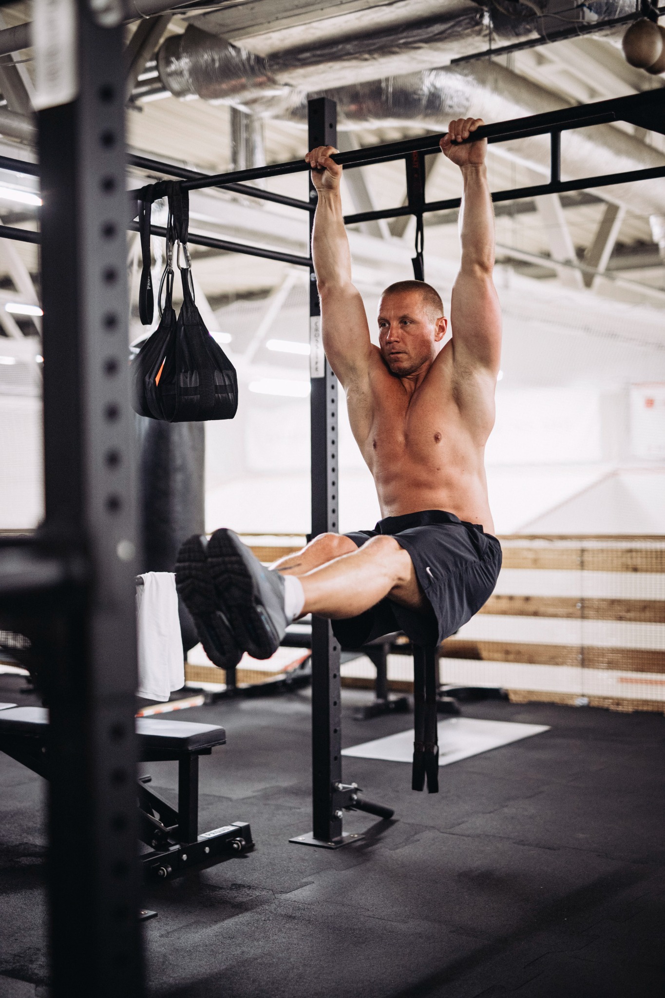 Mężczyzna w spodenkach i butach sportowych wykonuje ćwiczenie podciągania nóg na drążku w siłowni, z widocznym workiem treningowym i ławeczką w tle.