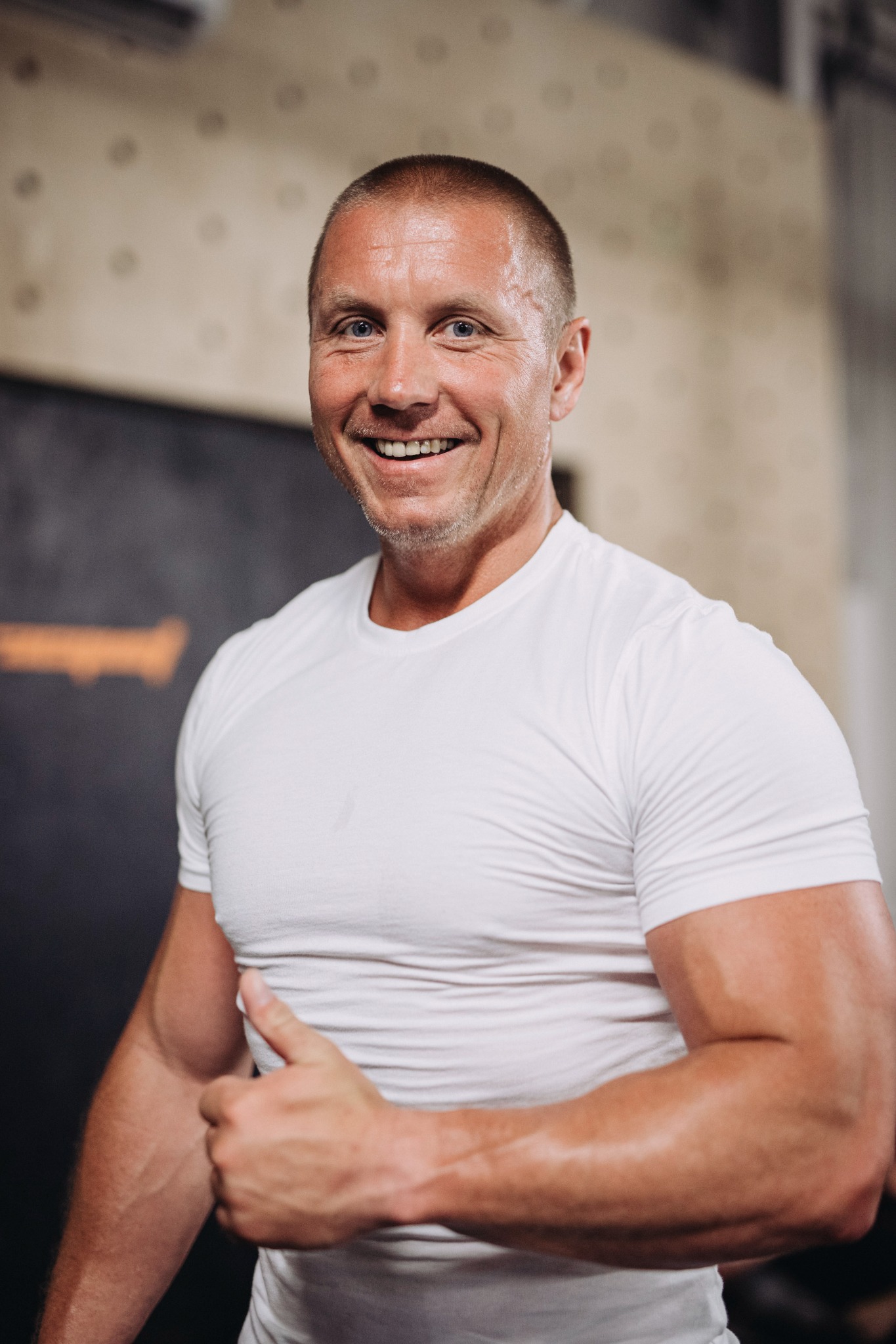 Uśmiechnięty mężczyzna w białej koszulce, z widocznymi mięśniami, pokazuje kciuk w górę na tle siłowni. Dobre samopoczucie i pozytywna energia.