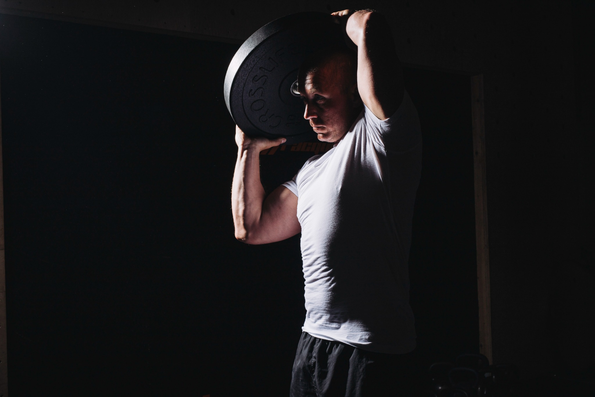 Mężczyzna w białej koszulce podnosi ciężar nad głową, oświetlony od dołu w ciemnym otoczeniu, prezentując siłę i determinację podczas treningu.