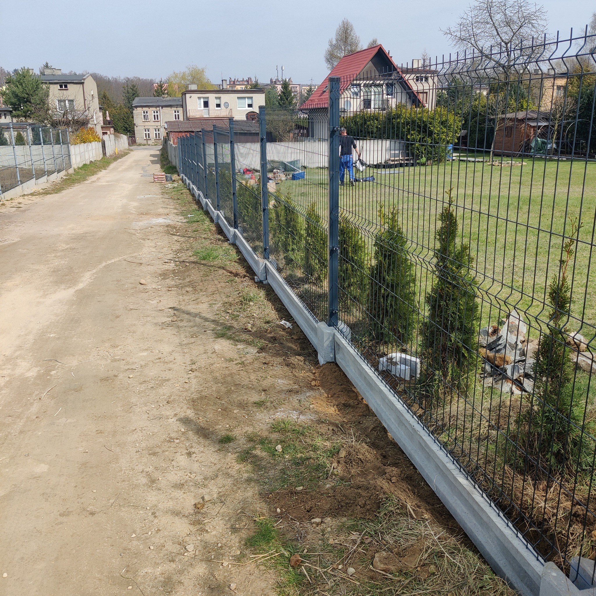 Długa linia nowo postawionego grafitowego ogrodzenia panelowego z betonową podmurówką, widok z boku drogi gruntowej, w tle zabudowania mieszkalne i osoba w niebieskich spodniach na posesji.