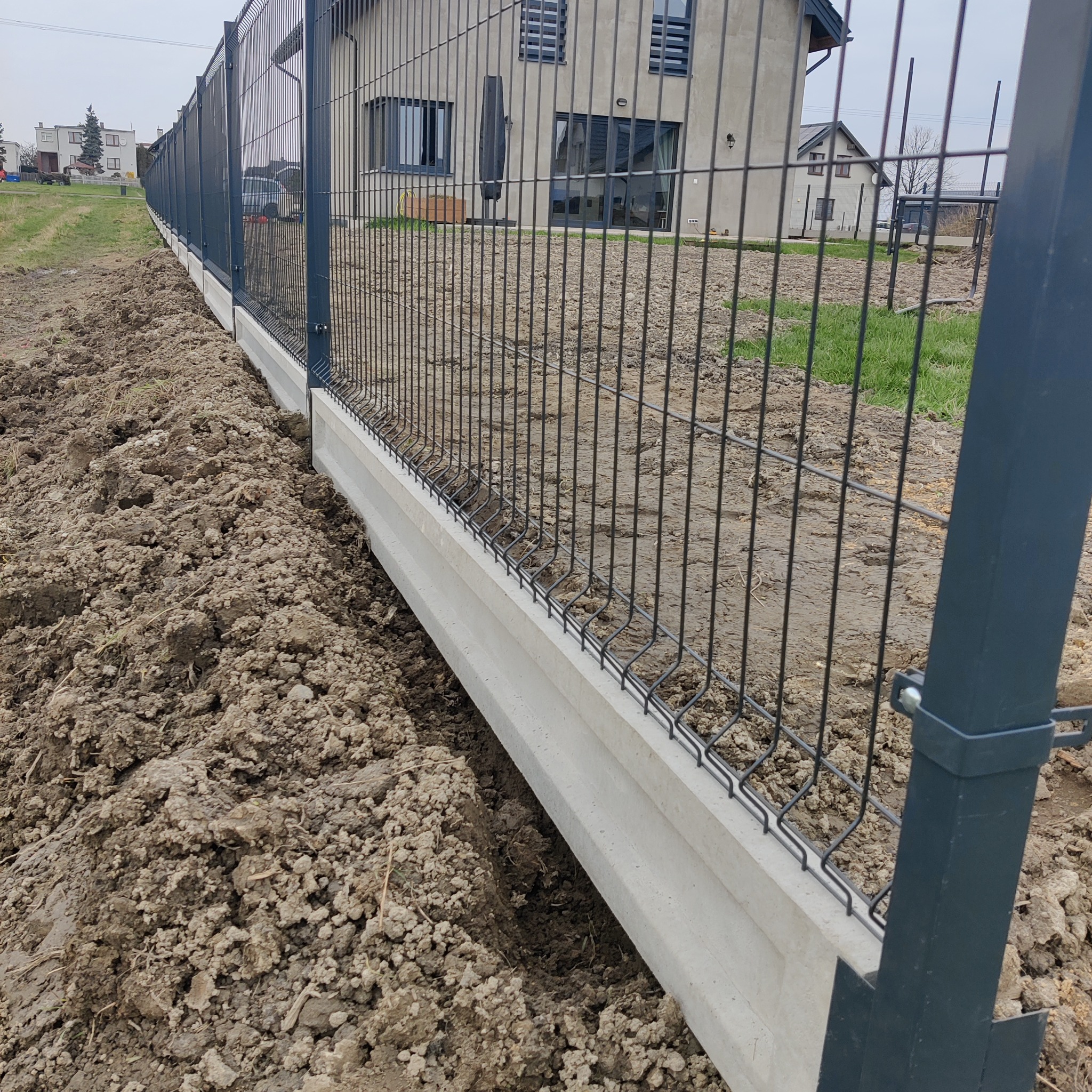 Nowo postawione, grafitowe ogrodzenie panelowe na betonowej podmurówce, otoczone świeżo przekopaną ziemią. W tle widoczne domy jednorodzinne.