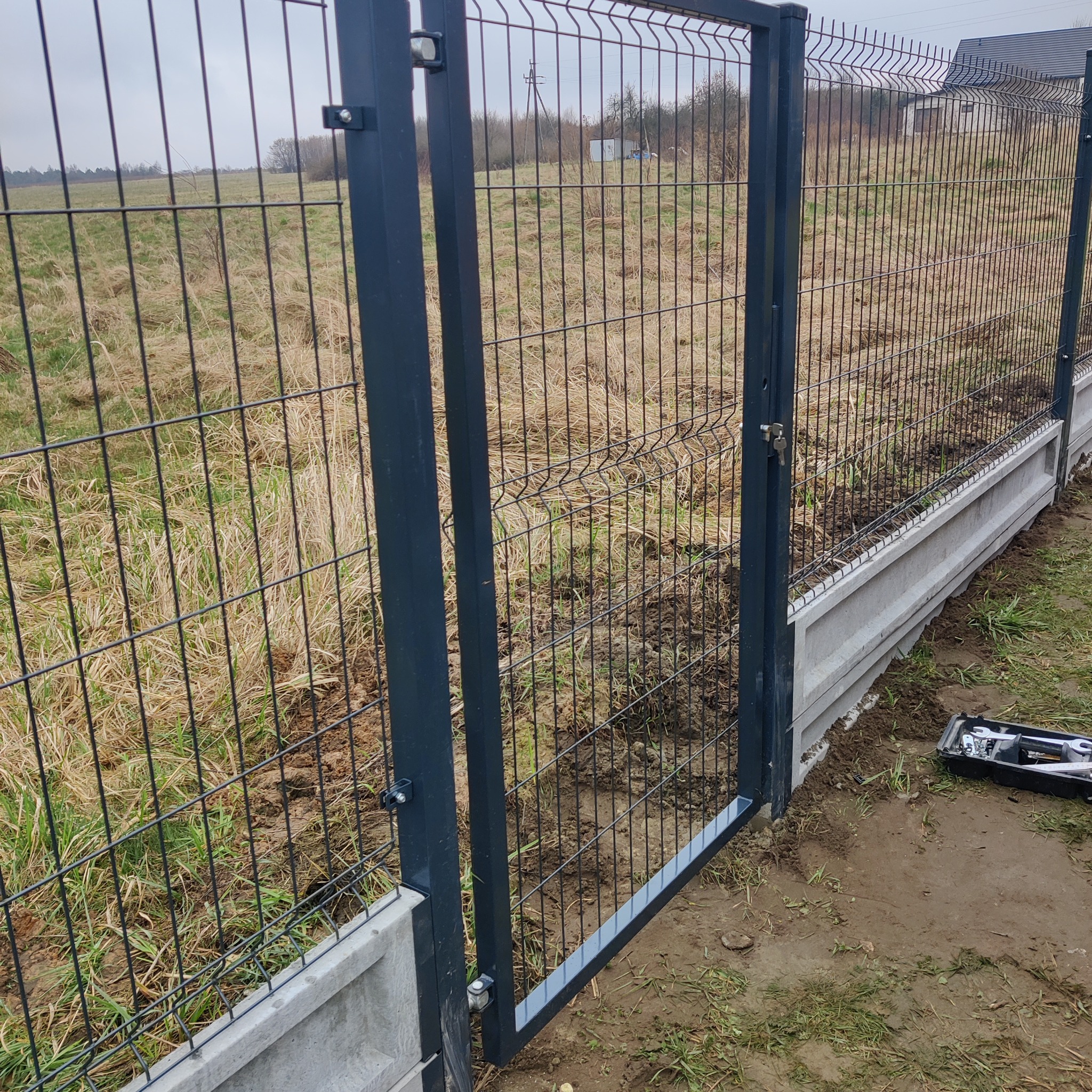 Nowo zamontowana brama wjazdowa z metalowej siatki na betonowej podmurówce, skrzynka z narzędziami obok, widok na pole w tle.