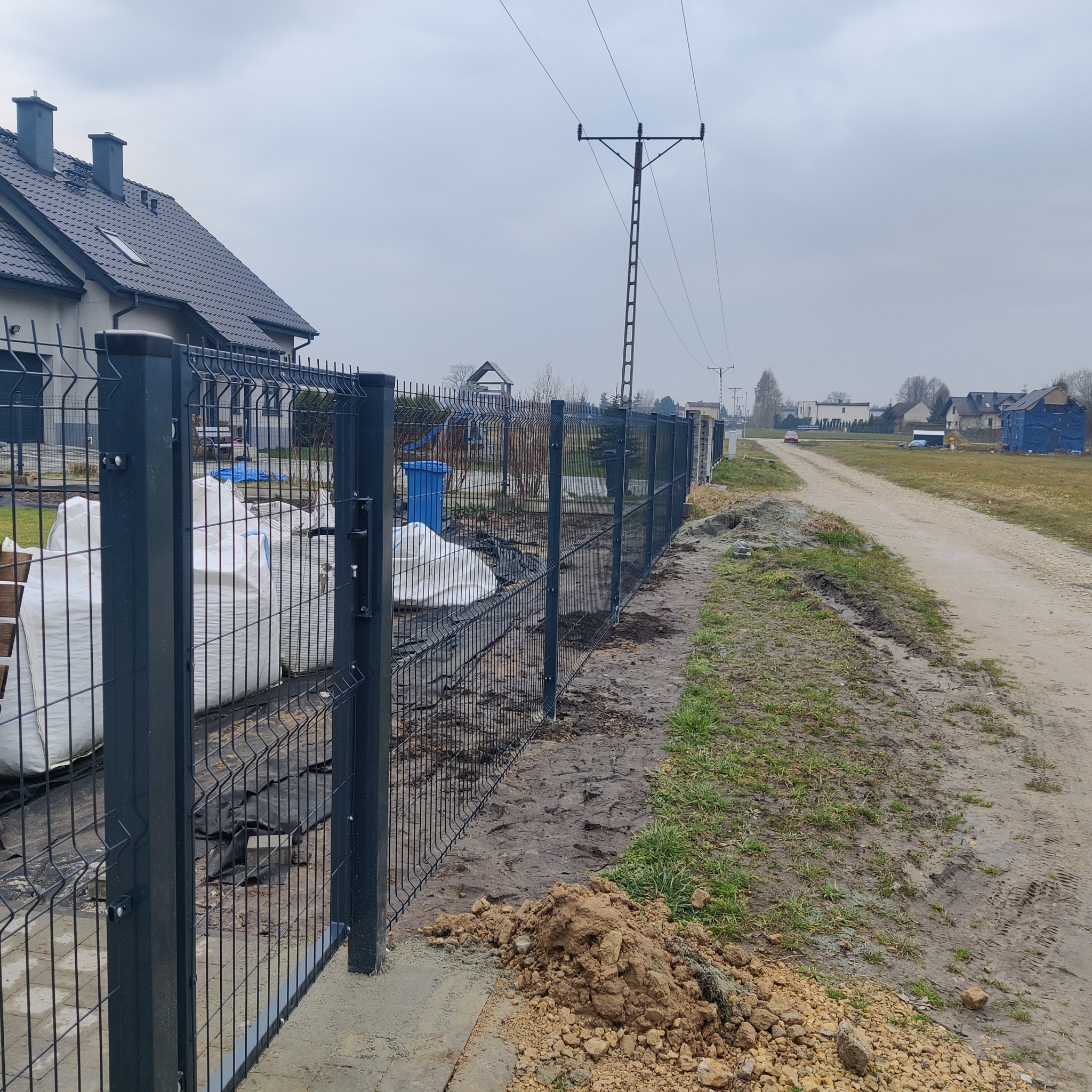 Nowoczesne grafitowe ogrodzenie panelowe na posesji z workami materiałów budowlanych w tle, widok z zewnątrz na drogę gruntową i słupy elektryczne.