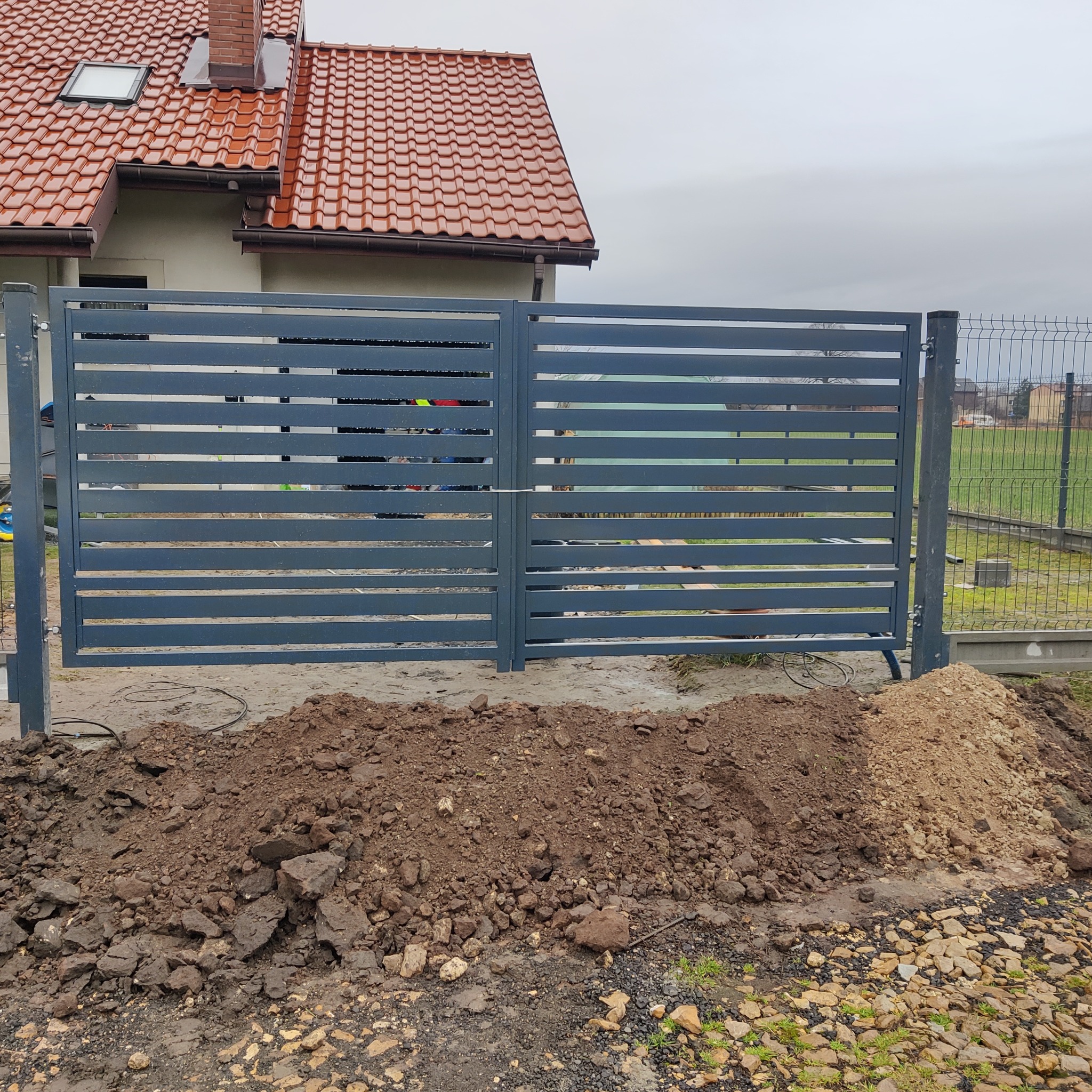 Nowoczesna, pozioma brama wjazdowa z metalu, szara, z widocznymi śladami prac ziemnych i stertą ziemi u podstawy, na tle domu z czerwoną dachówką i siatki ogrodzeniowej.