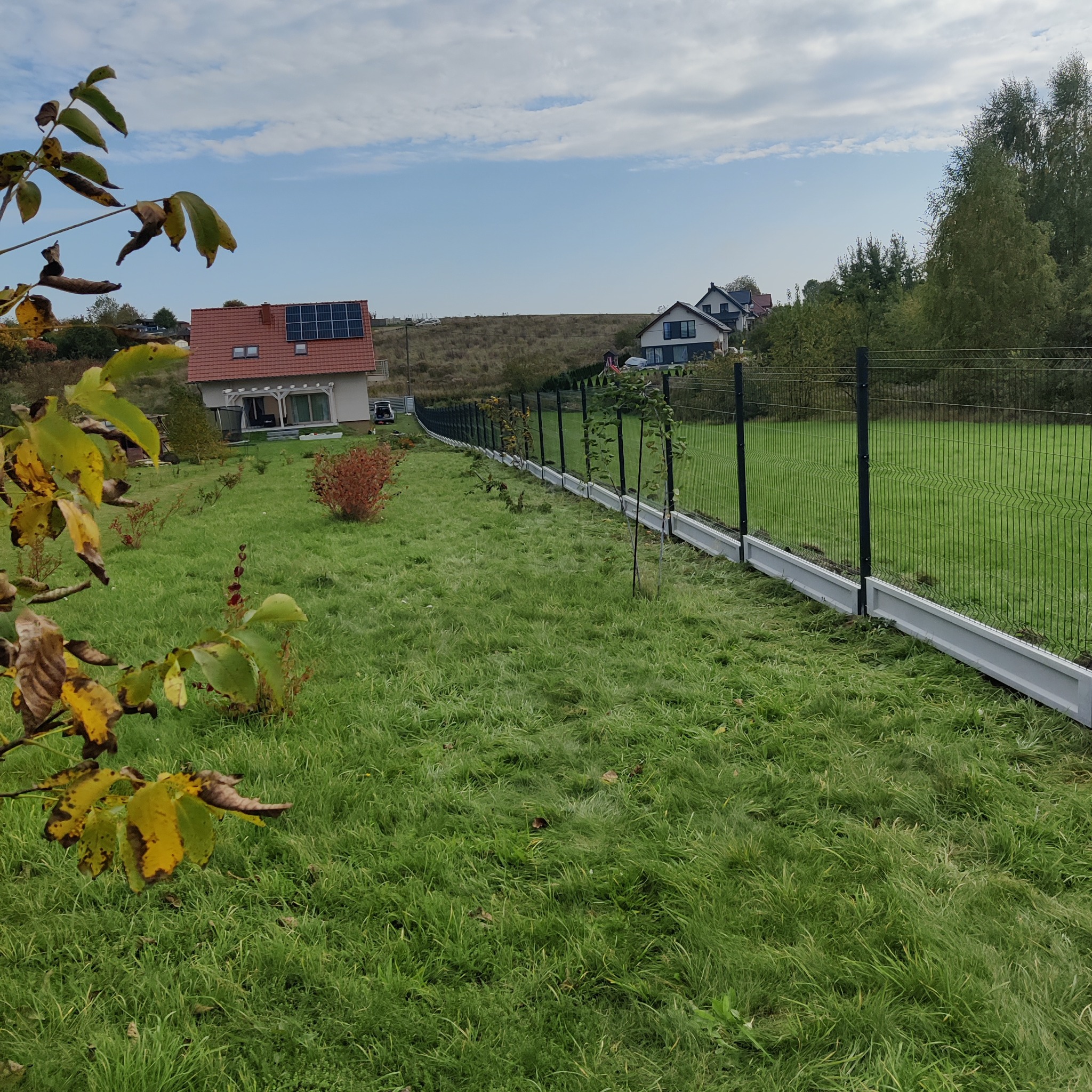 Długa linia czarnego ogrodzenia panelowego z szarą podmurówką biegnąca wzdłuż trawnika, z widokiem na domy w tle pod błękitnym niebem z chmurami.