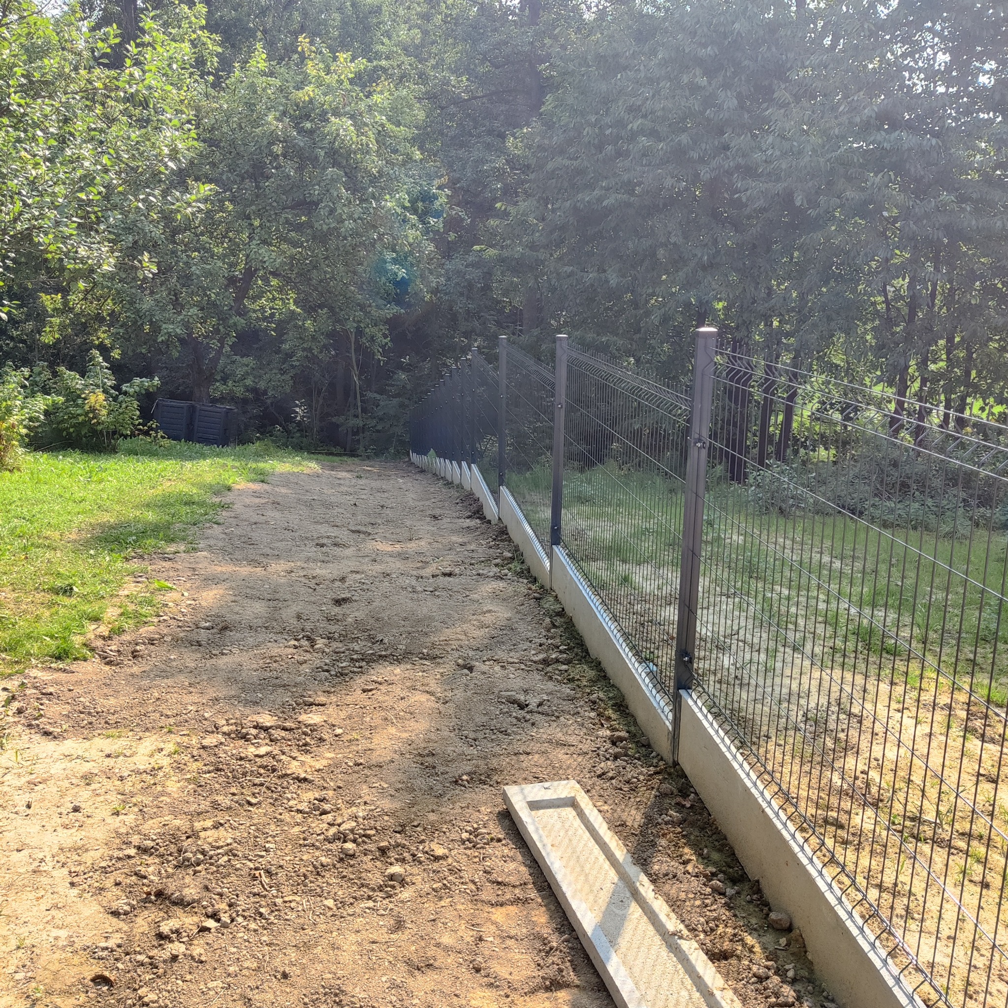 Długi fragment nowego ogrodzenia z siatki z podmurówką betonową, biegnący wzdłuż ścieżki z ubitej ziemi, w tle drzewa.