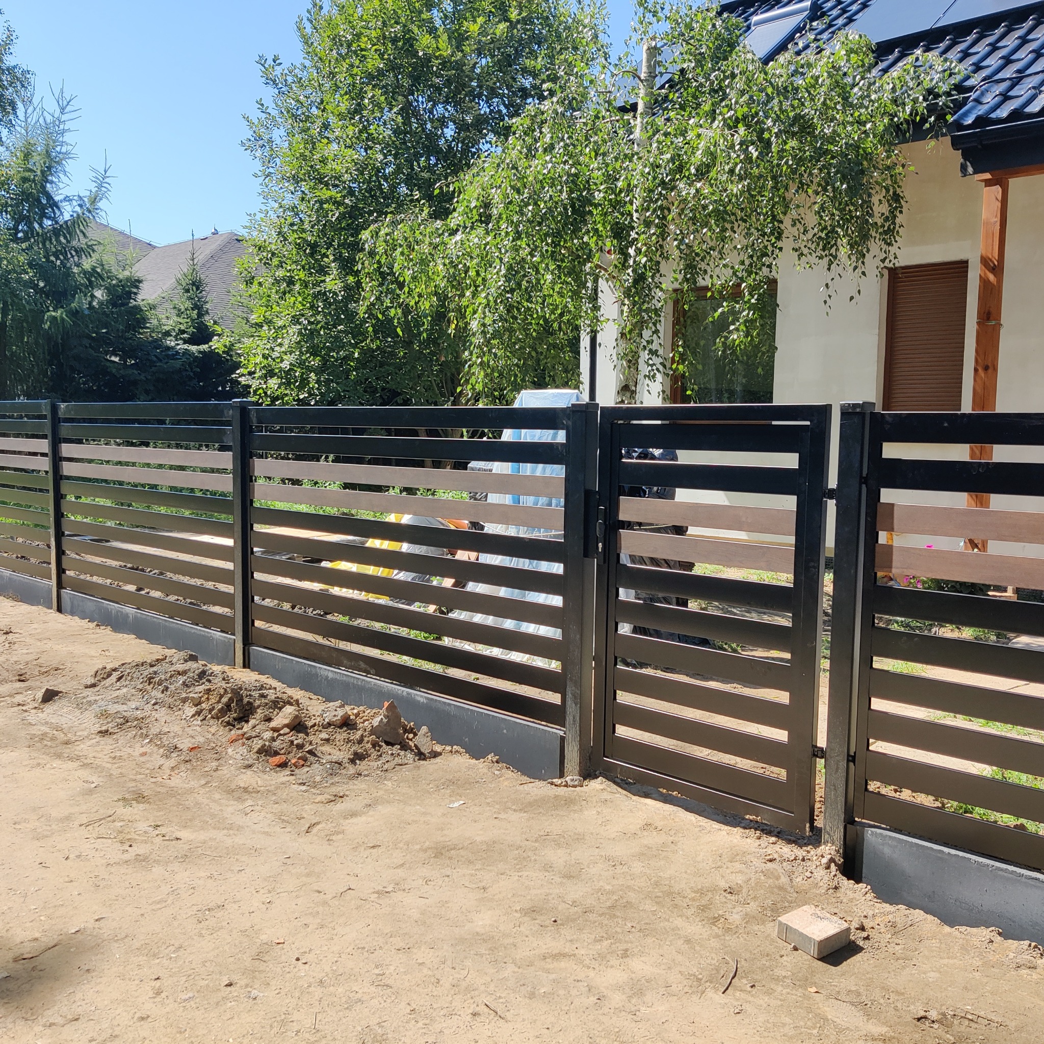 Nowoczesne ogrodzenie posesji z poziomymi panelami w kolorze brązowym i czarnym, widoczne prace ziemne.