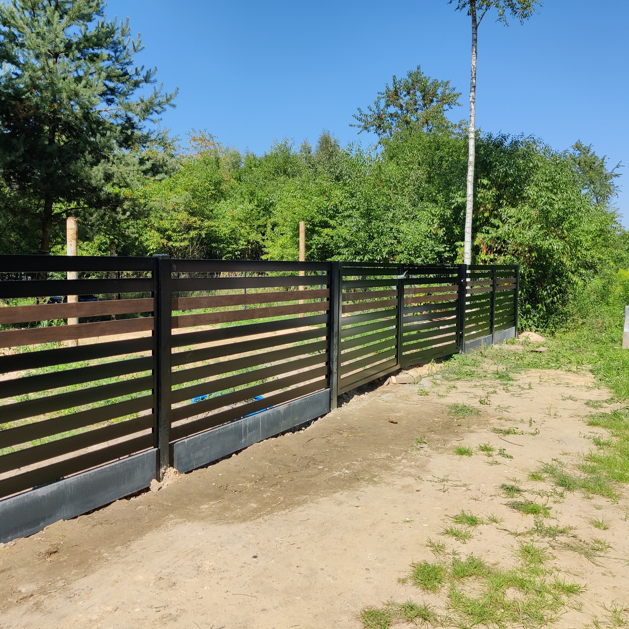 Nowoczesne ogrodzenie z poziomych paneli w kolorze drewna i czarnych słupków, z widocznymi betonowymi podmurówkami, na tle zieleni i błękitnego nieba.