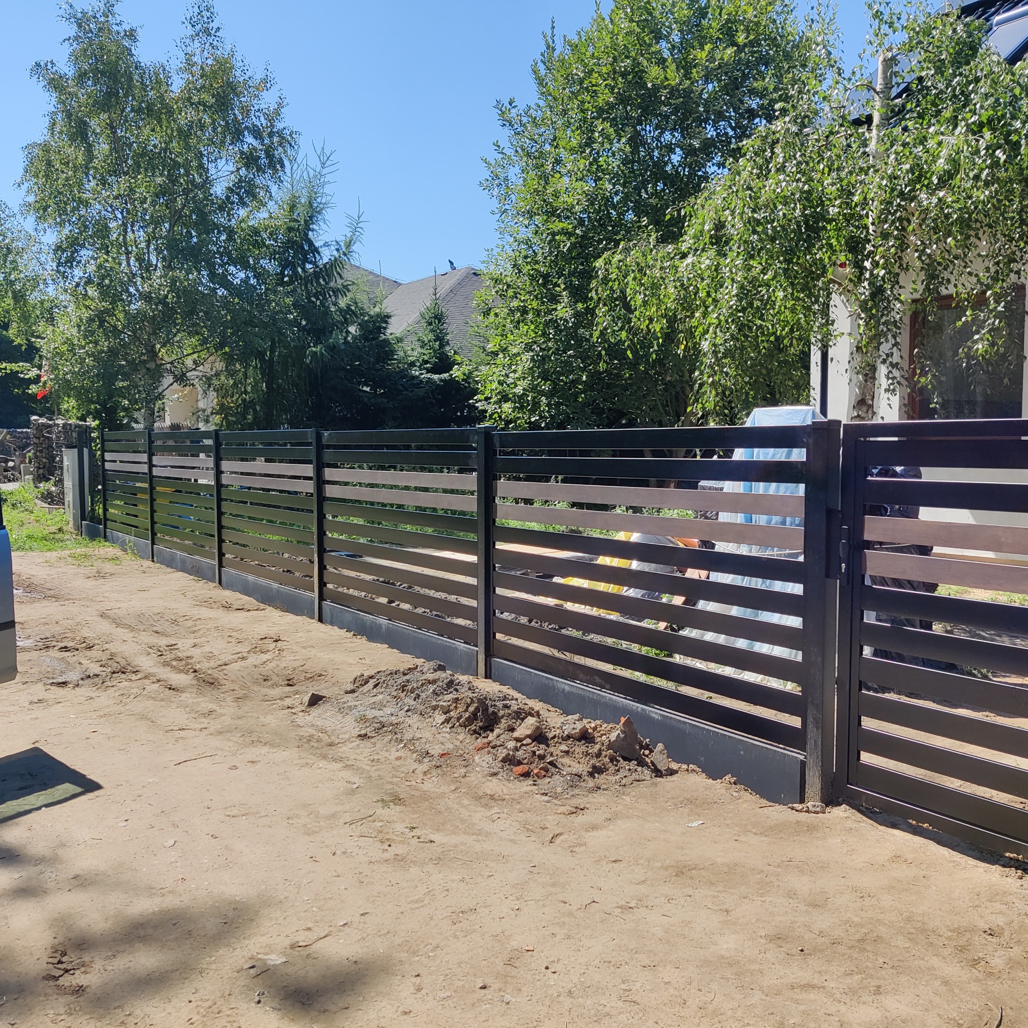 Nowoczesne, poziome ogrodzenie z metalowych profili w kolorze antracytowym, z bramą wjazdową, widoczne ślady prac ziemnych.