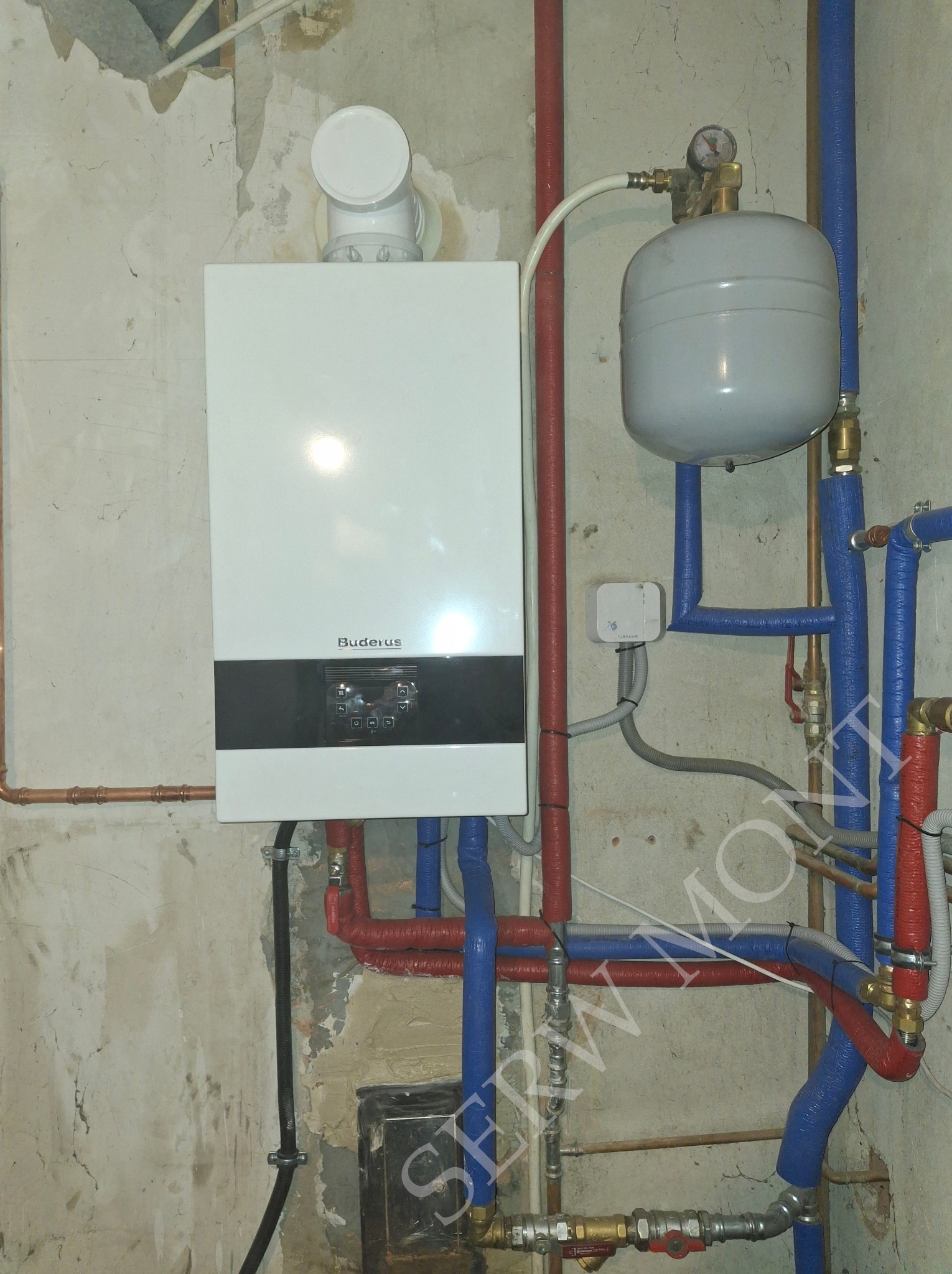 Zainstalowany jednofunkcyjny kocioł gazowy Buderus Gb122i z orurowaniem w kolorach czerwonym i niebieskim, zamontowany na tle surowej, nieotynkowanej ściany z widocznymi śladami wcześniejszych...