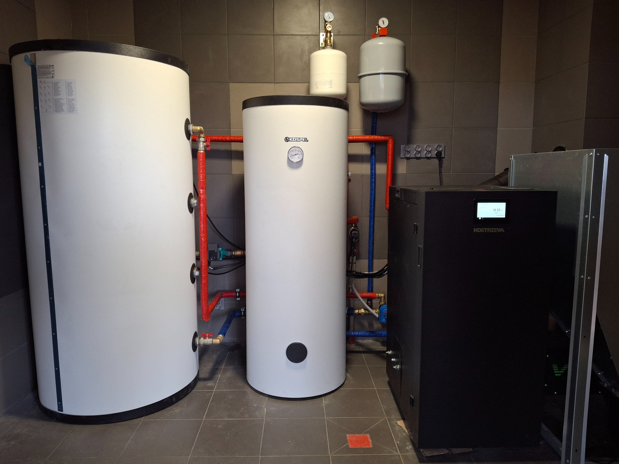 Dwa białe zbiorniki akumulacyjne ciepła, czarny piec z wyświetlaczem, oraz białe zbiorniki wyrównawcze na tle szarych płytek, połączone rurami w kolorach czerwonym i niebieskim.