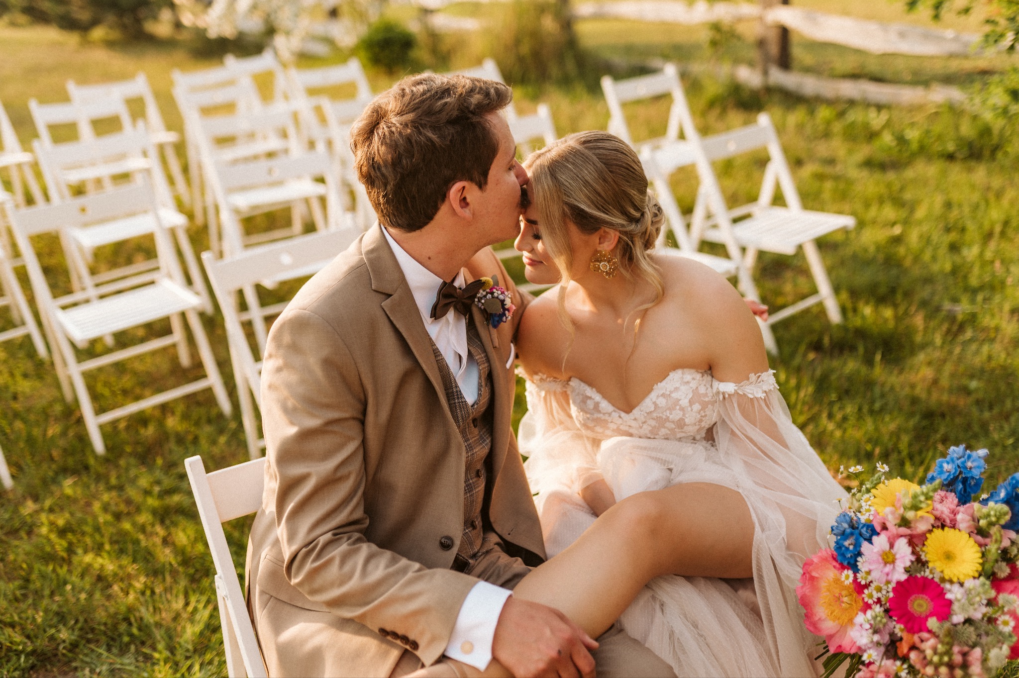 Para młoda siedząca na krzesłach w plenerze, mężczyzna w beżowym garniturze całuje kobietę w sukni ślubnej w czoło, w tle rząd białych krzeseł i kwiaty.