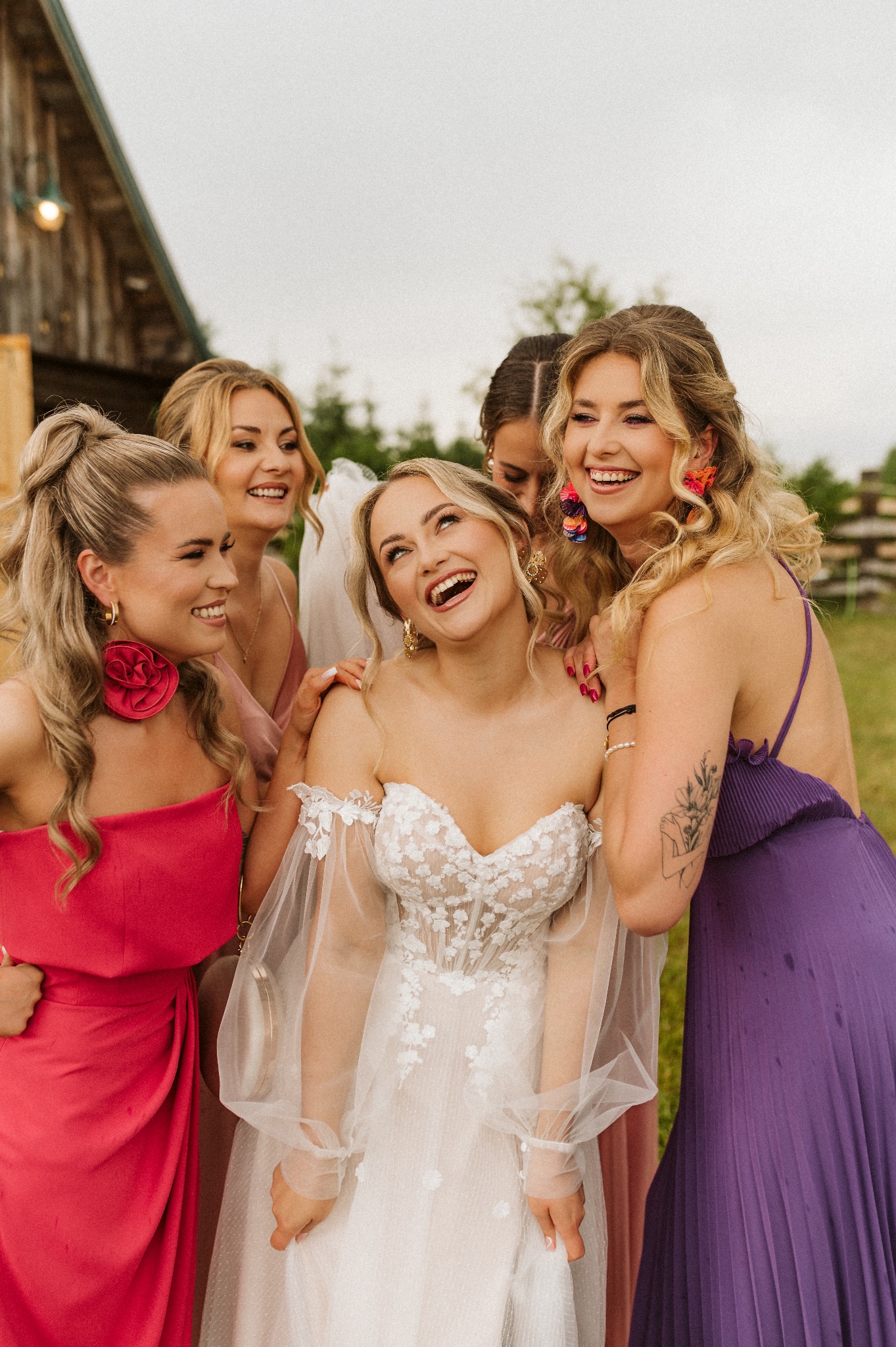 Uśmiechnięta panna młoda w koronkowej sukni otoczona czterema druhnami w kolorowych sukienkach, w tle drewniany budynek i zieleń.