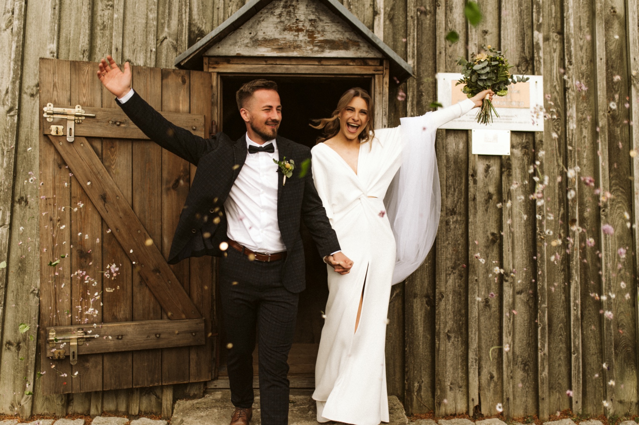 Uśmiechnięta para młoda wychodzi z drewnianej stodoły podczas ceremonii obsypywania kwiatami, panna młoda w długiej białej sukni z rozcięciem, pan młody w ciemnym garniturze w kratę i muszce.