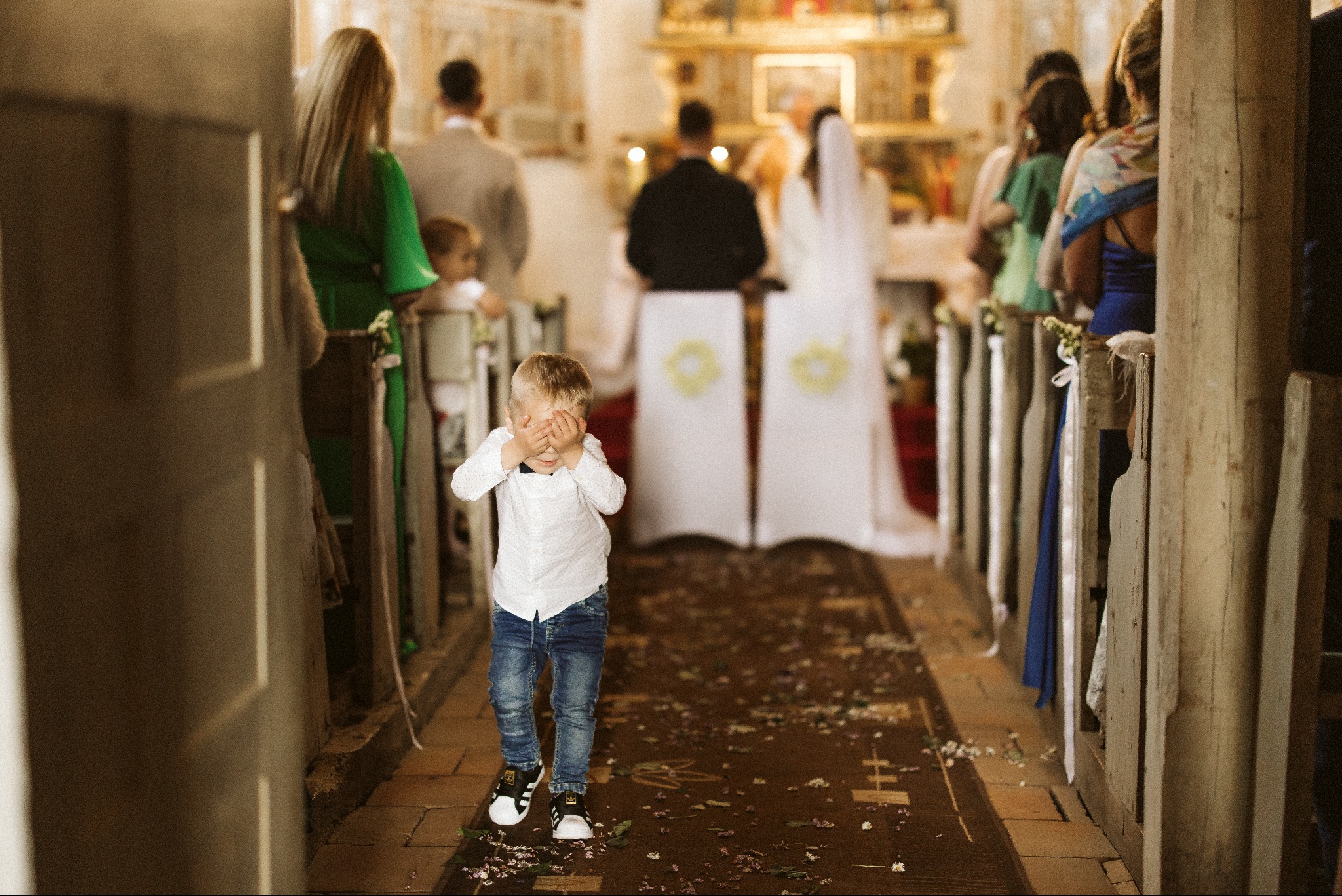 Chłopiec w koszuli i jeansach zakrywa oczy na tle pary młodej podczas ceremonii ślubnej w kościele, z rozsypanymi kwiatami na dywanie.